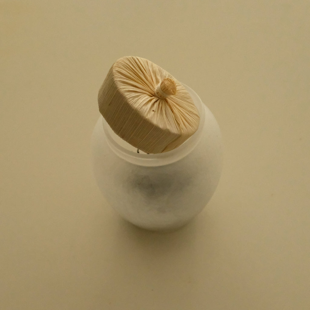 A small white round container with a translucent lid, topped with a beige fabric pouch secured by a string, set against a plain beige background.