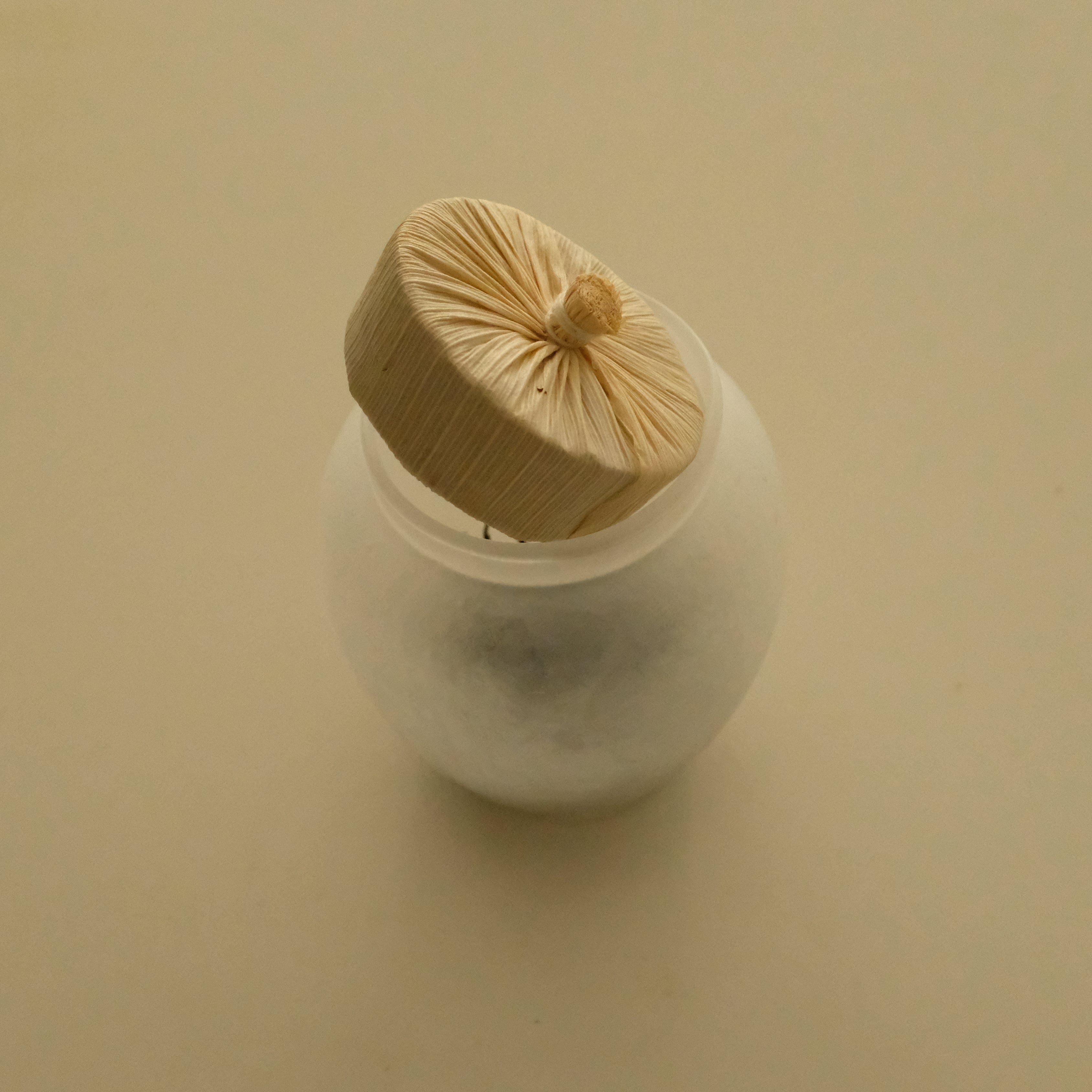 A small white round container with a translucent lid, topped with a beige fabric pouch secured by a string, set against a plain beige background.