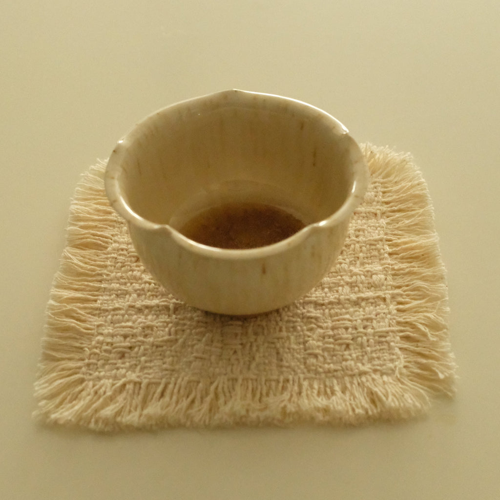 A small handmade ceramic cup with a scalloped rim sits on a square woven fringed coaster against a solid cream background.