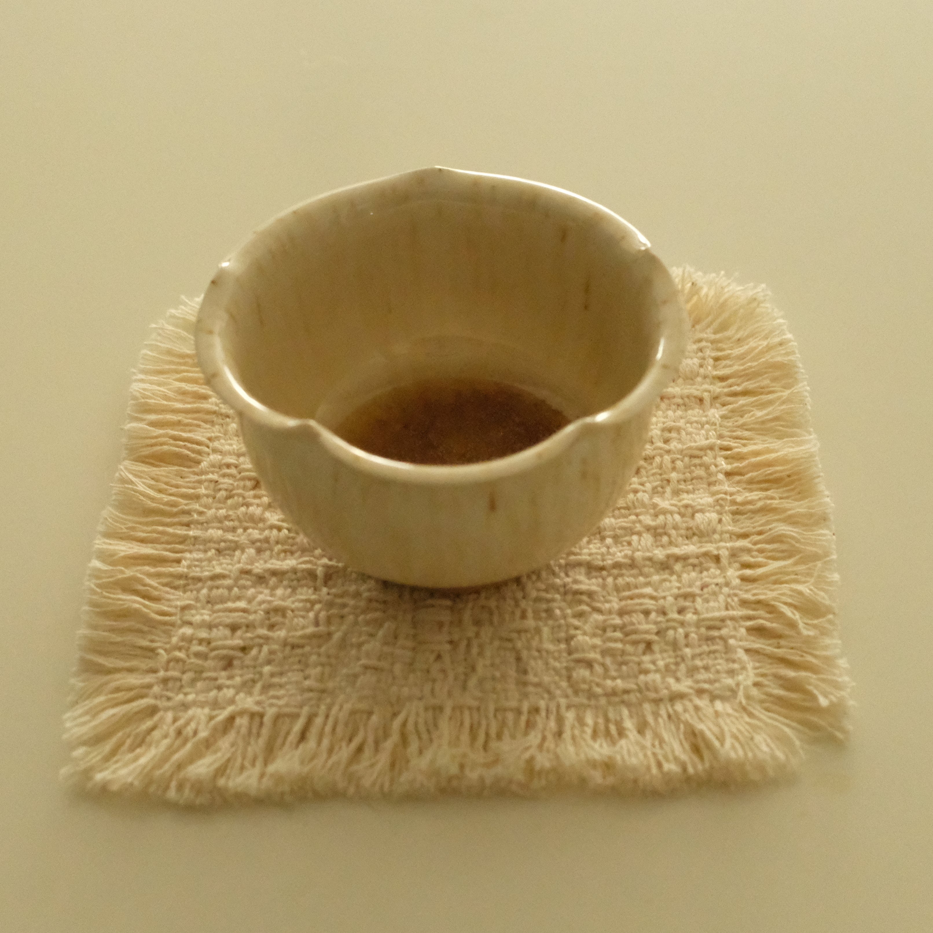 A small handmade ceramic cup with a scalloped rim sits on a square woven fringed coaster against a solid cream background.