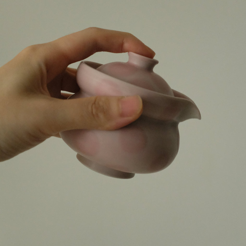 A hand holding a small pink ceramic teapot with a lid and spout against a plain light gray background.