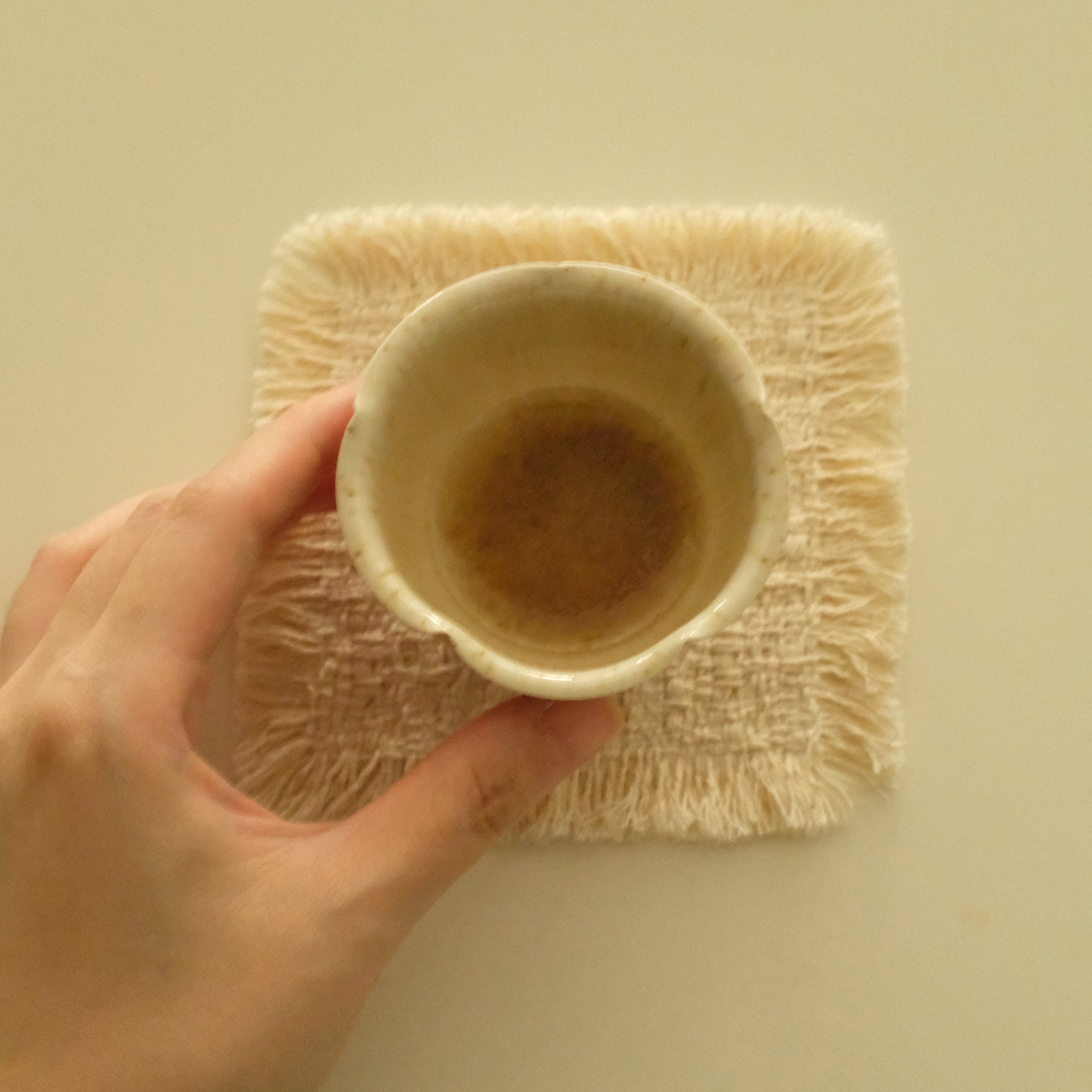 A hand holding a small beige ceramic cup over a square cream-colored textured fringed mat on a plain beige background.