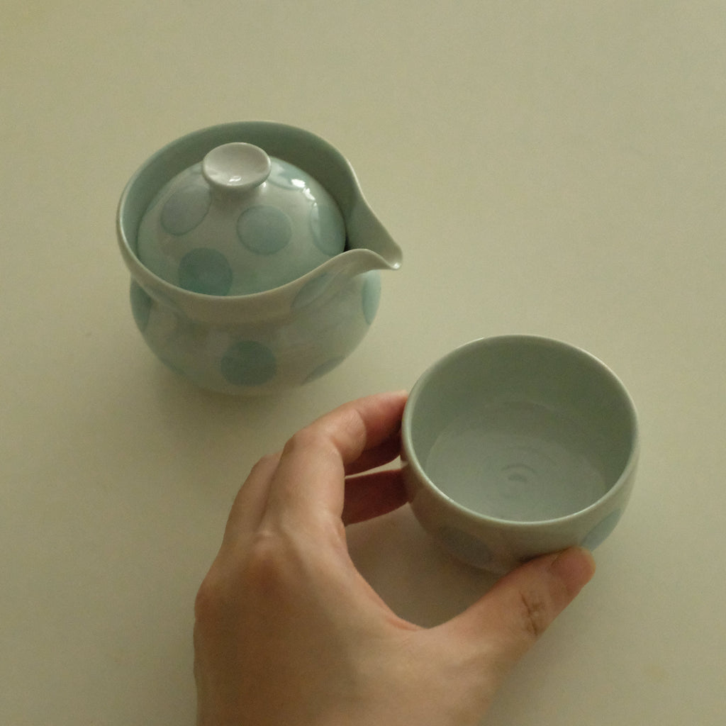 A hand holds a small pale blue-green ceramic cup next to a matching lidded teapot with large polka dots on a plain light beige surface.