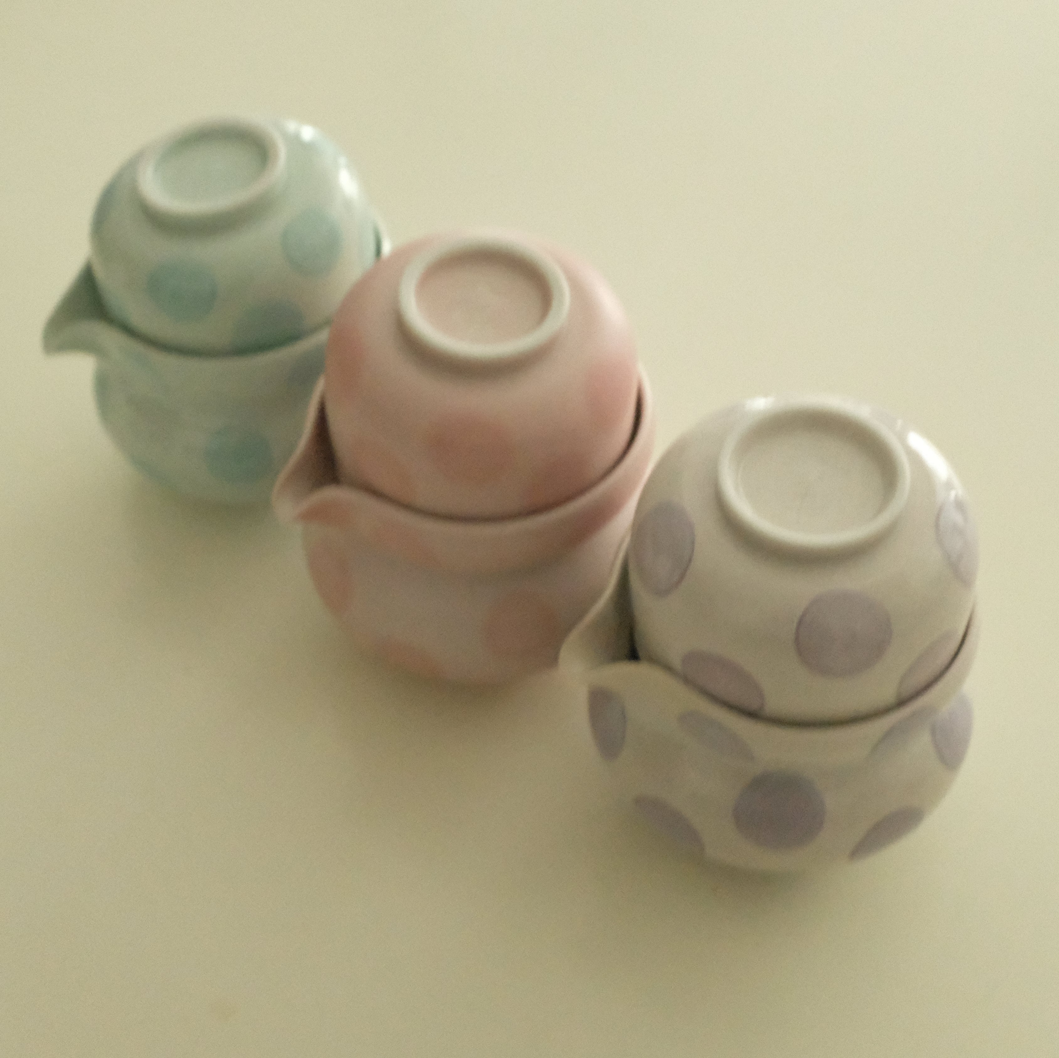 Three small stacked ceramic teapot-like containers with lids, decorated with polka dots in pastel mint green, pink, and lavender on a plain light beige background.