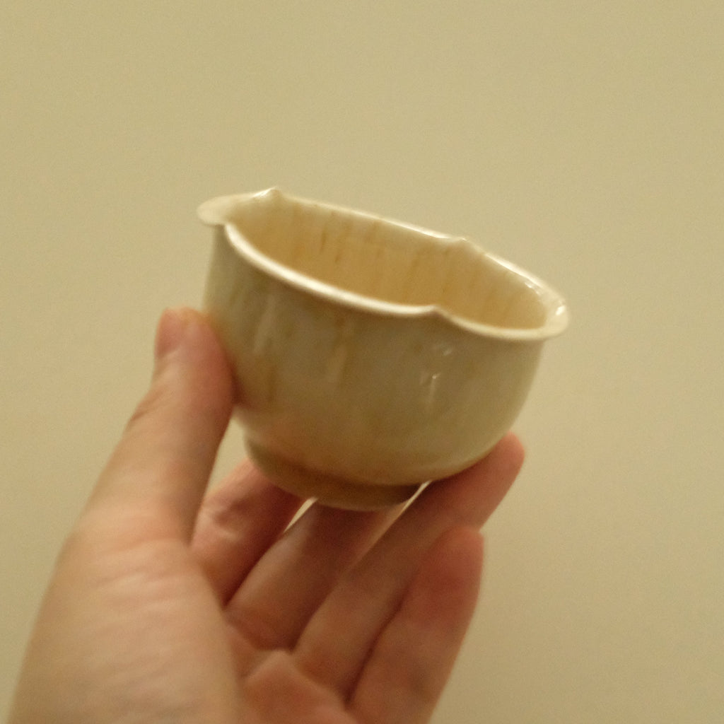 A hand holding a small handmade ceramic cup with a wide, shallow bowl and a scalloped rim, shown against a plain beige background.
