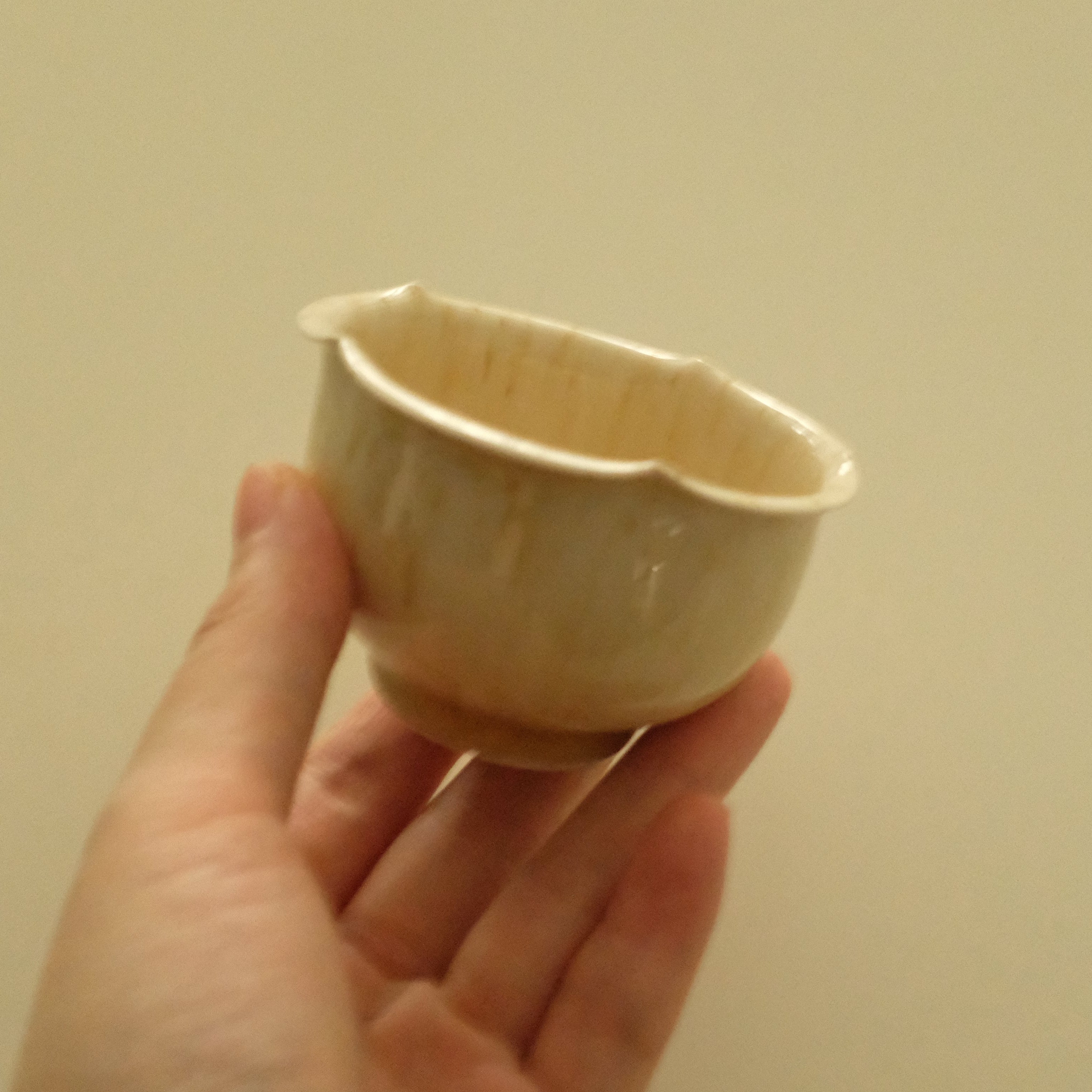A hand holding a small handmade ceramic cup with a wide, shallow bowl and a scalloped rim, shown against a plain beige background.