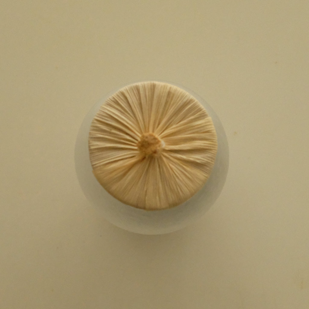 A round, light beige textured item with a gathered center, resting on a small white circular base against a solid cream-colored background.