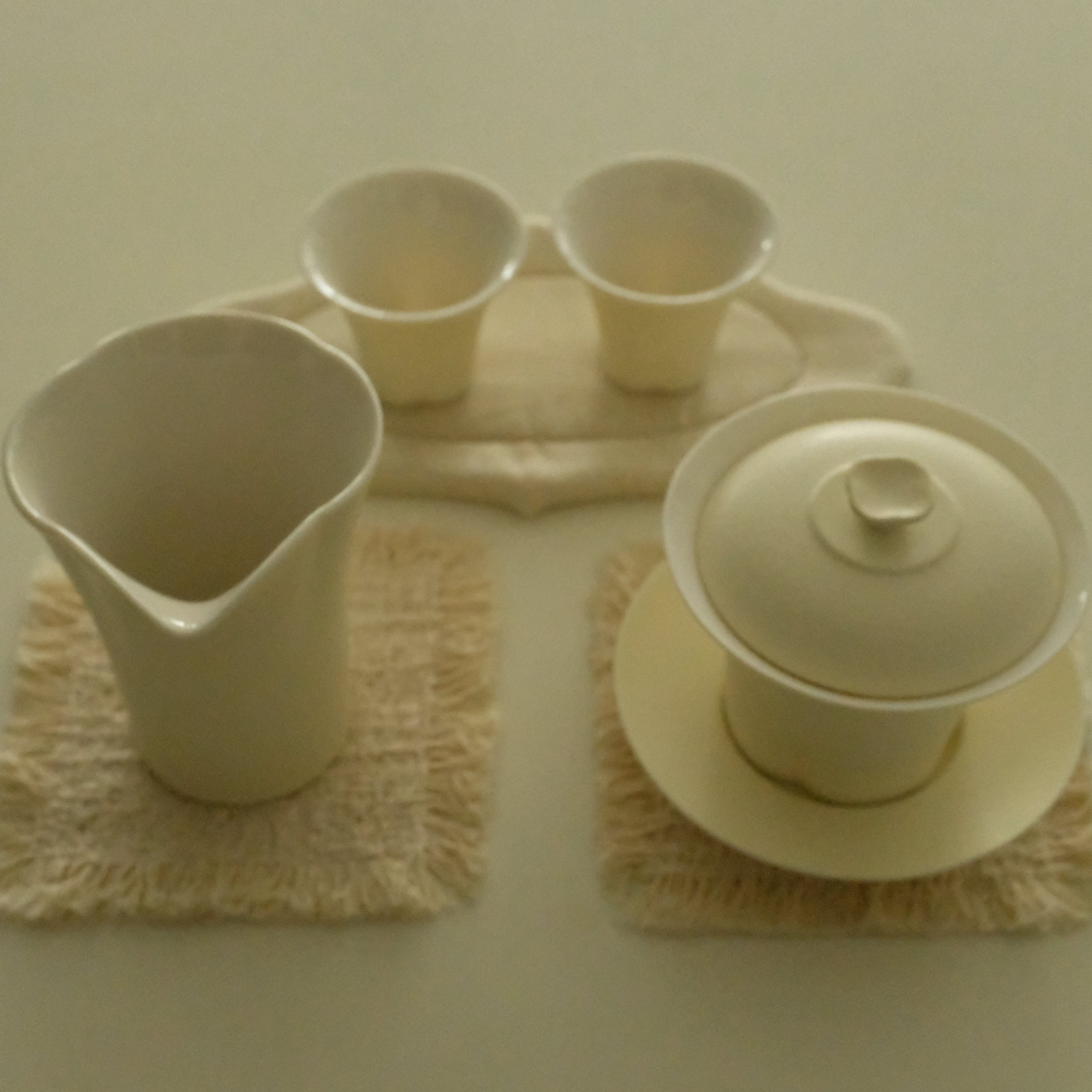 A ceramic tea set in a warm cream or off-white glaze, including a lidded teapot, a pitcher, two small cups, and matching saucers, arranged on textured woven mats against a plain light background.