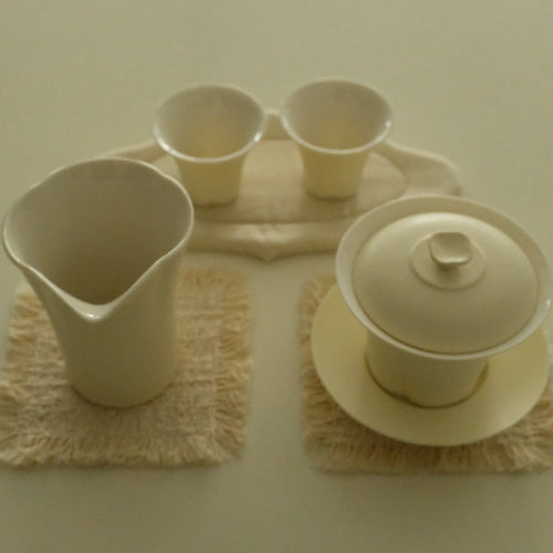 A ceramic tea set in a warm cream or off-white glaze, including a lidded teapot, a pitcher, two small cups, and matching saucers, arranged on textured woven mats against a plain light background.