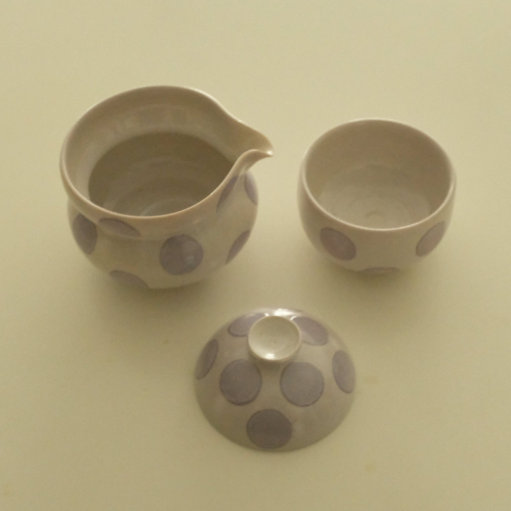 A three-piece ceramic tea set with a small handless teapot, a small cup, and a matching lid, all decorated with large lavender polka dots on a light beige background.