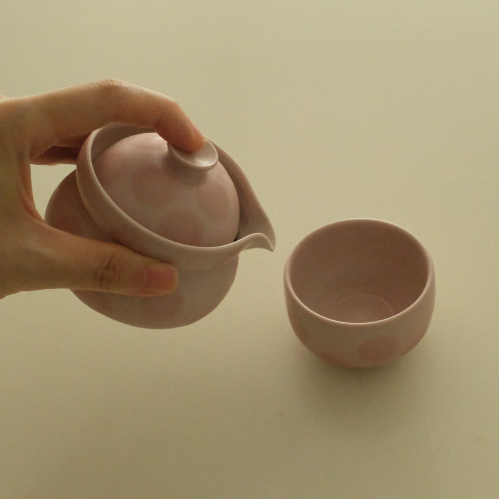 A hand holds a small pink ceramic teapot with a lid and spout above a matching pink cup on a plain light beige background.