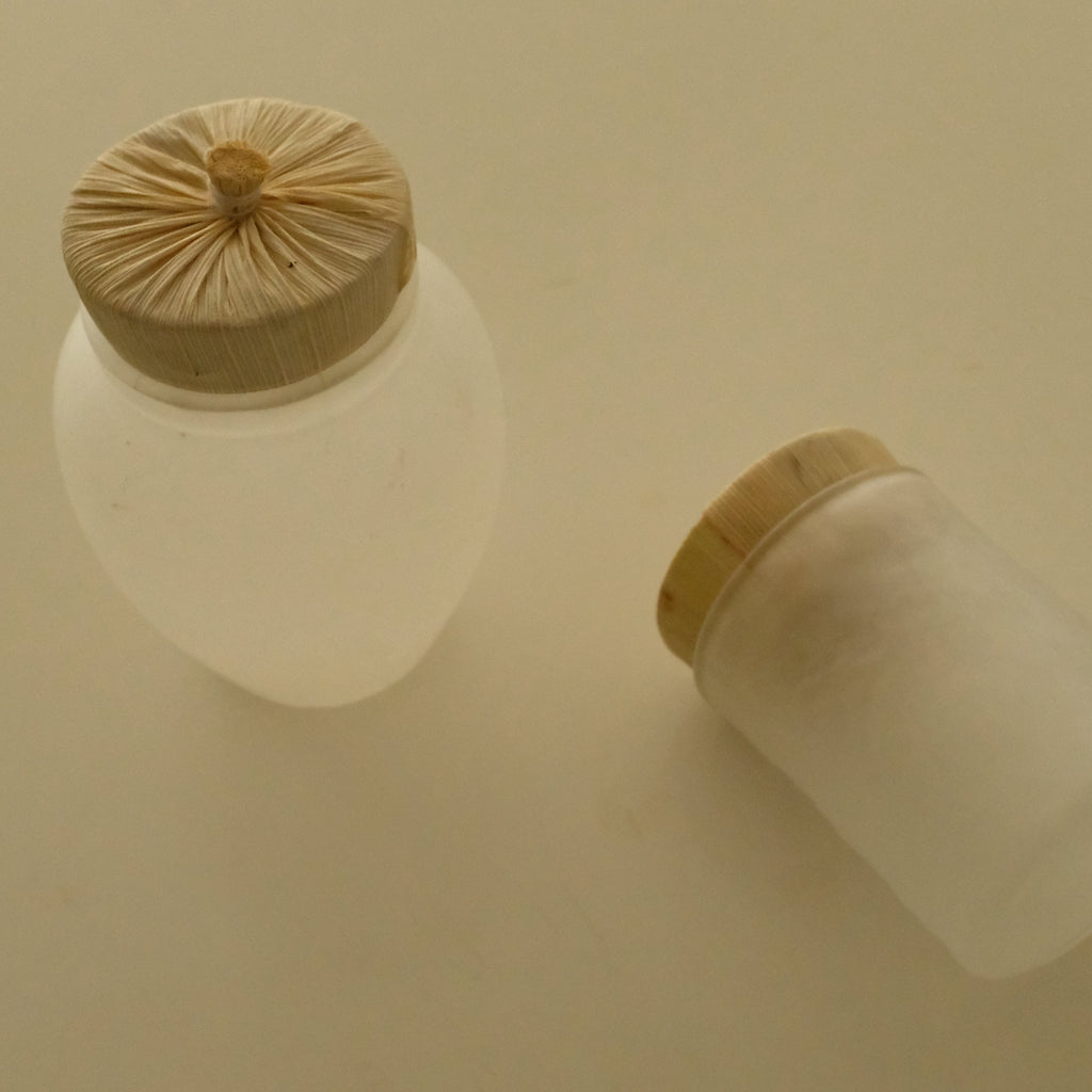 Two frosted glass jars with light wood lids on a warm beige background, one standing upright and the other lying on its side.