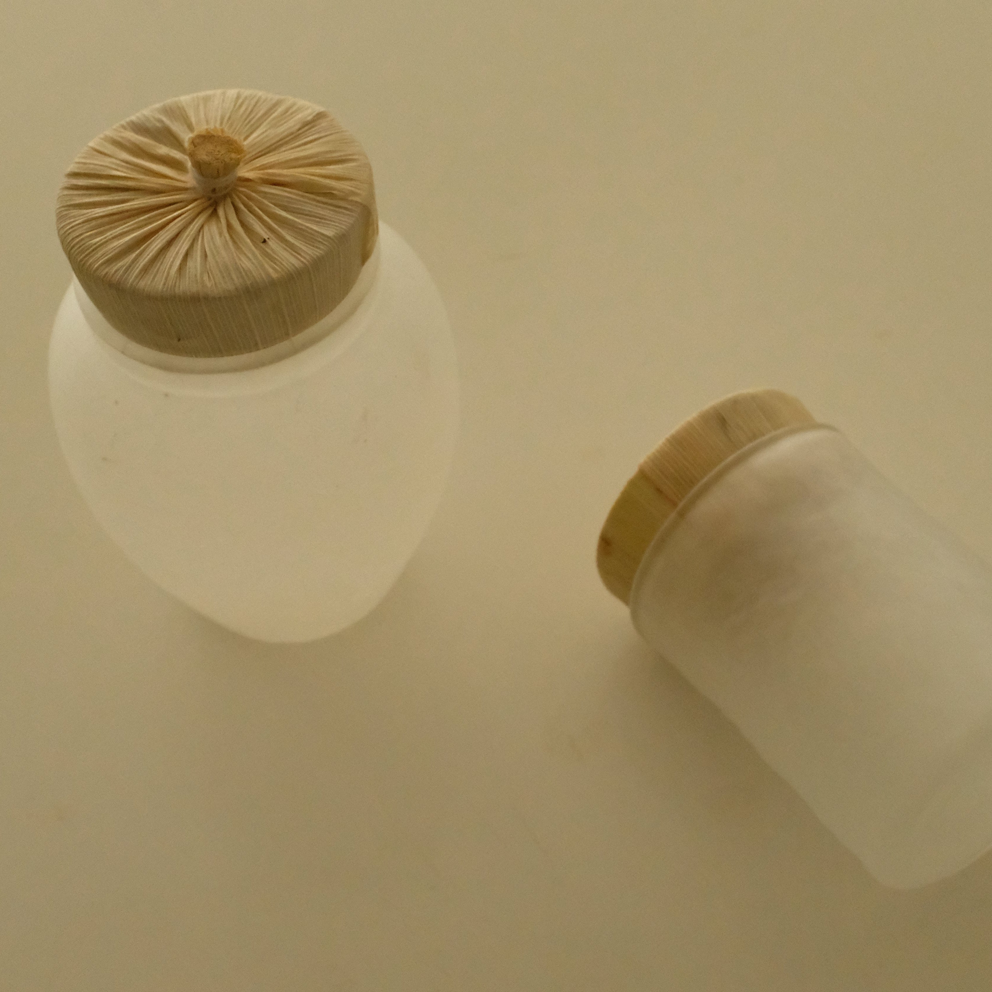 Two frosted glass jars with light wood lids on a warm beige background, one standing upright and the other lying on its side.