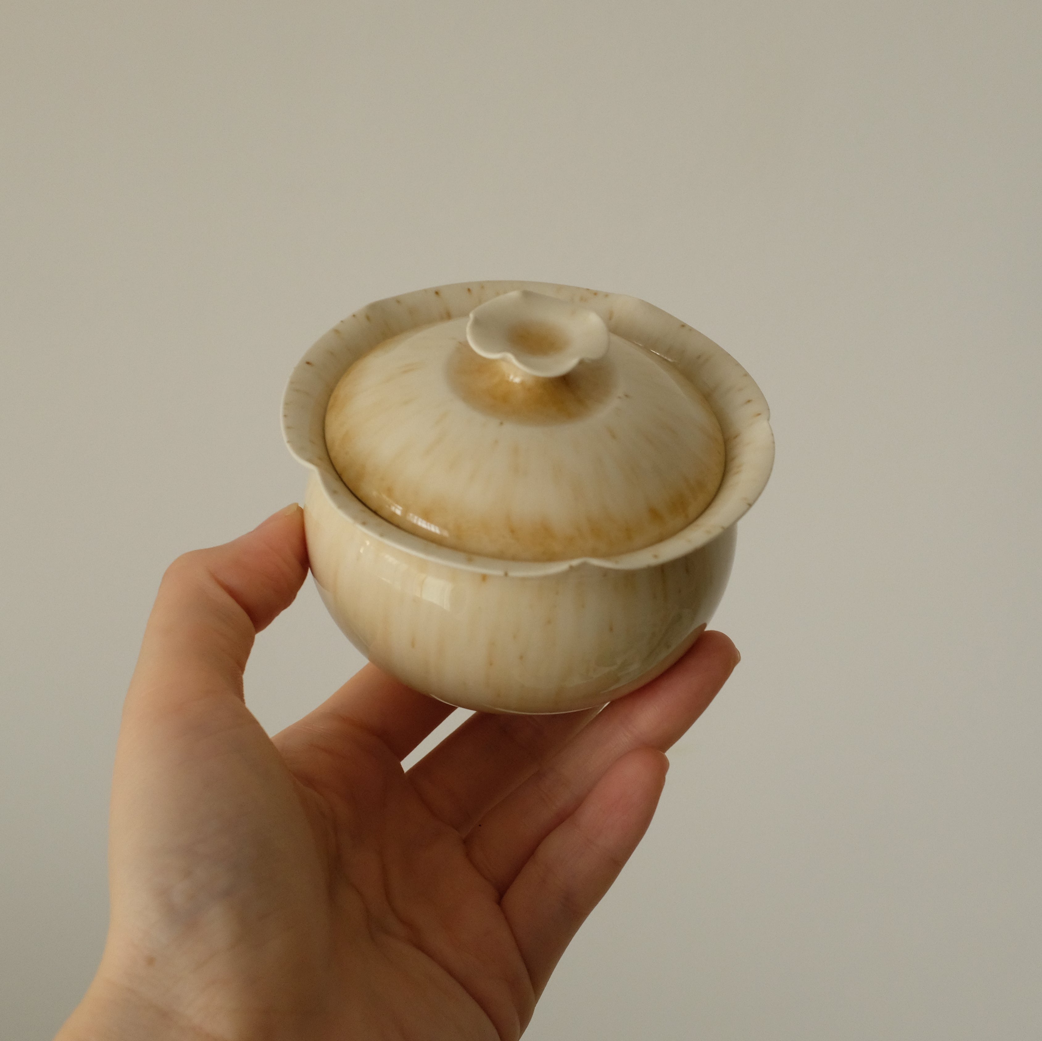 A hand holding a small ceramic lidded cup or teacup with a glossy cream and light brown glaze against a plain light gray background.
