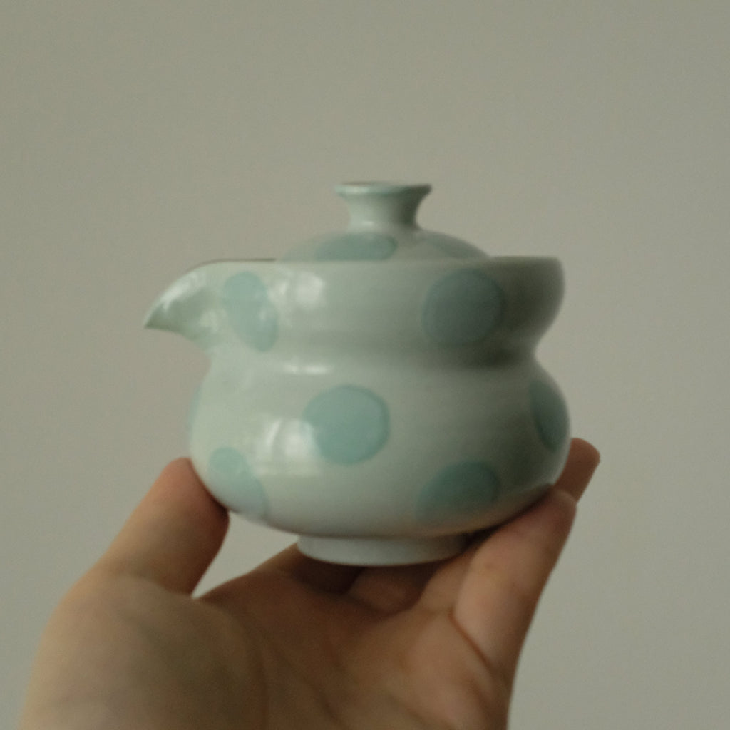 A hand holding a small glossy ceramic teapot with a pale green glaze and light blue polka dots against a plain light gray background.