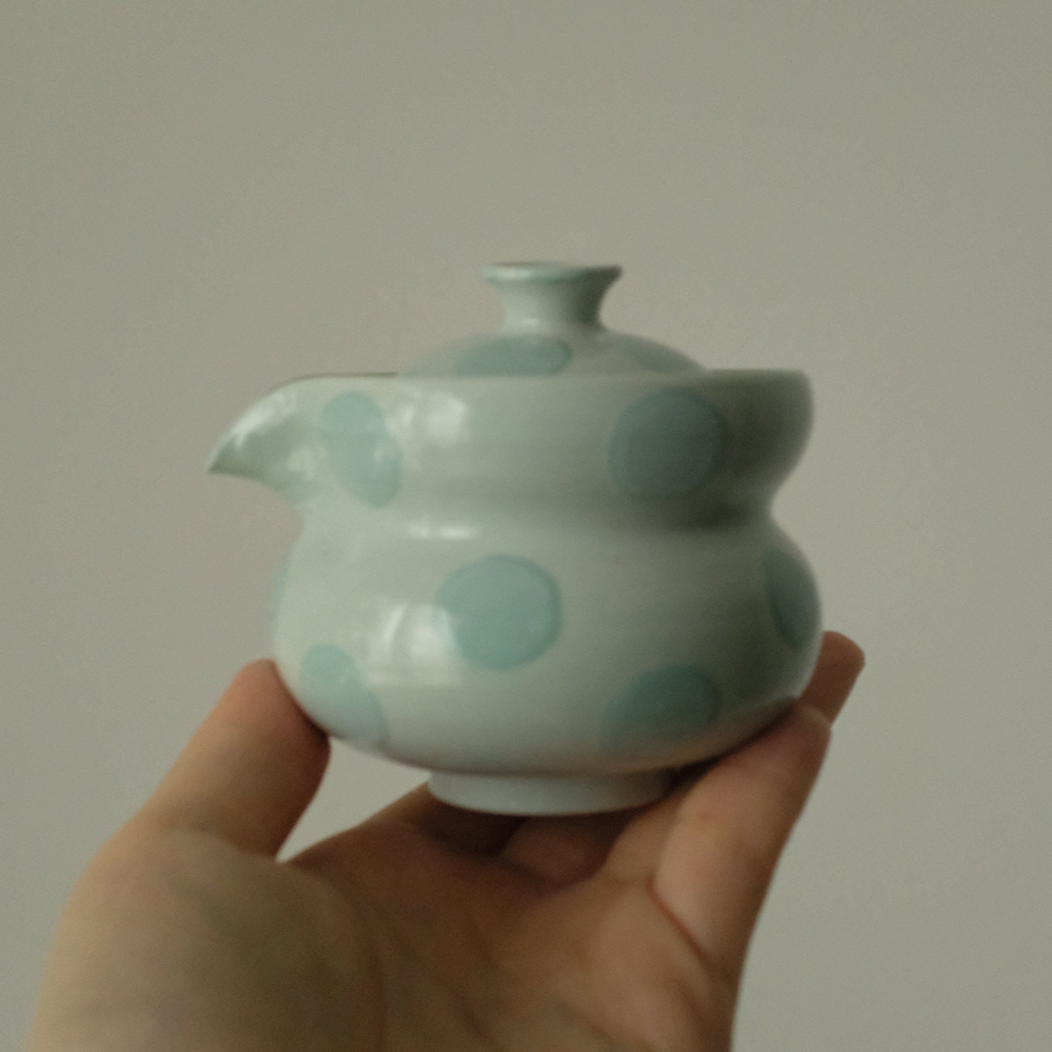 A hand holding a small glossy ceramic teapot with a pale green glaze and light blue polka dots against a plain light gray background.