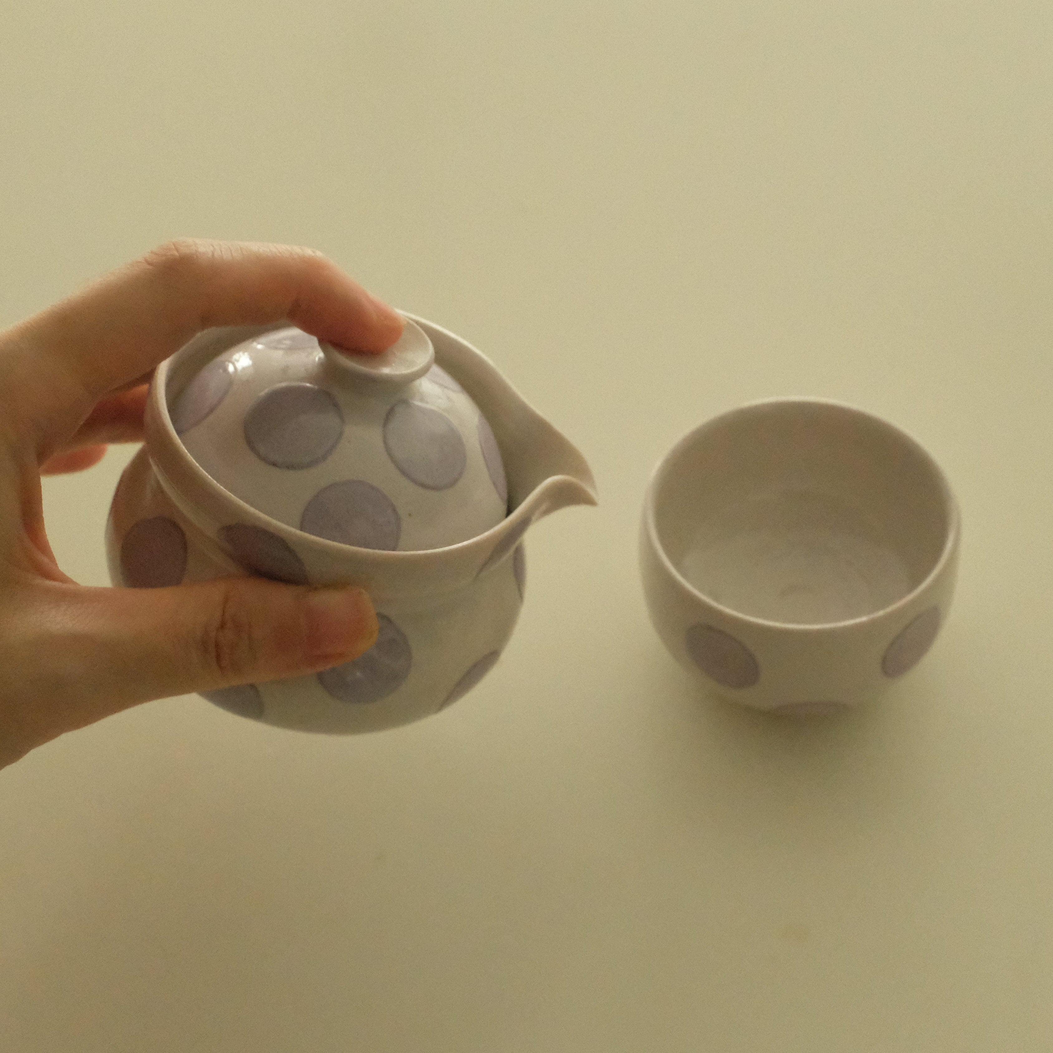 A hand holds a small ceramic handless teapot with a lid and spout, decorated with large gray polka dots on a white glaze, next to a matching small cup on a plain light background.