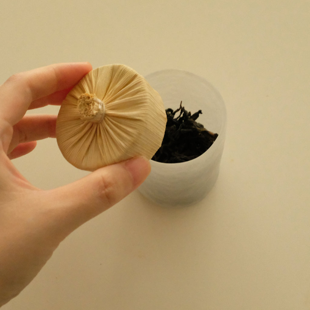 A hand holding a small beige fabric-wrapped round object in front of a clear plastic cup containing dark dried plant material, on a plain light beige background.