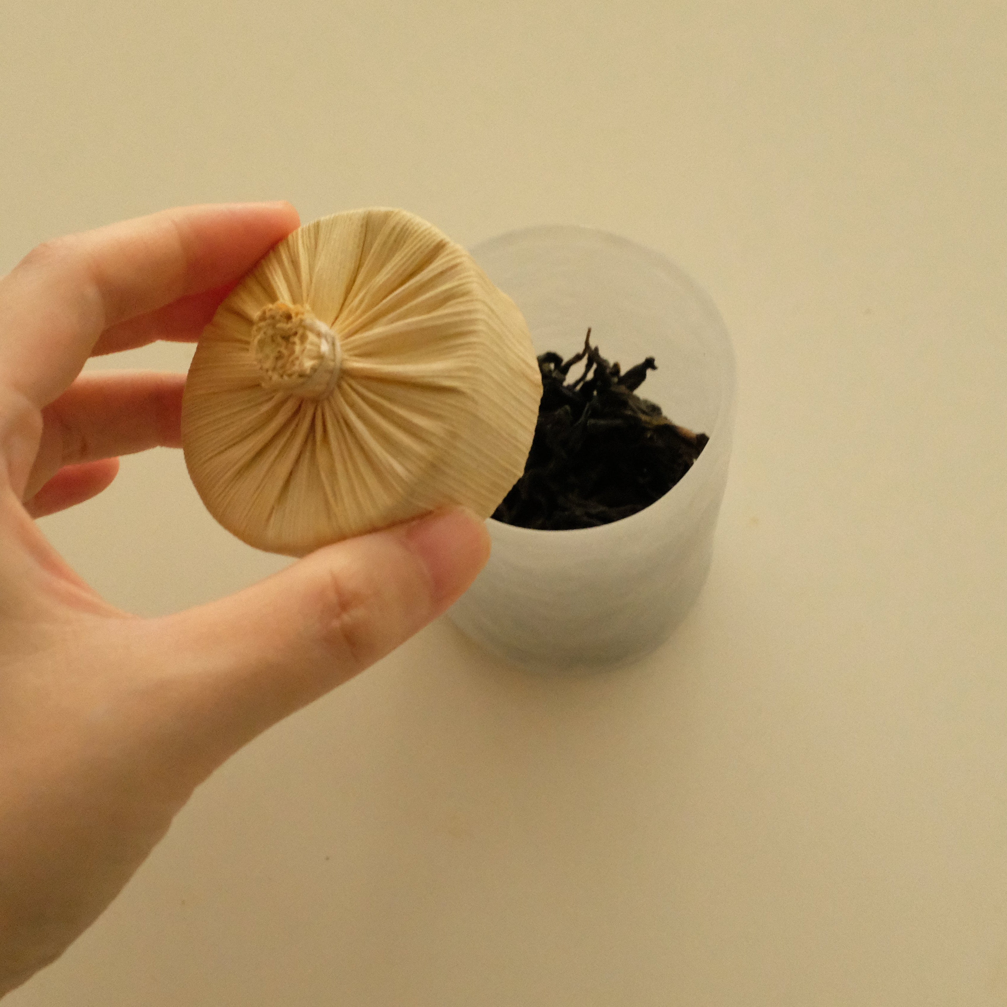 A hand holding a small beige fabric-wrapped round object in front of a clear plastic cup containing dark dried plant material, on a plain light beige background.