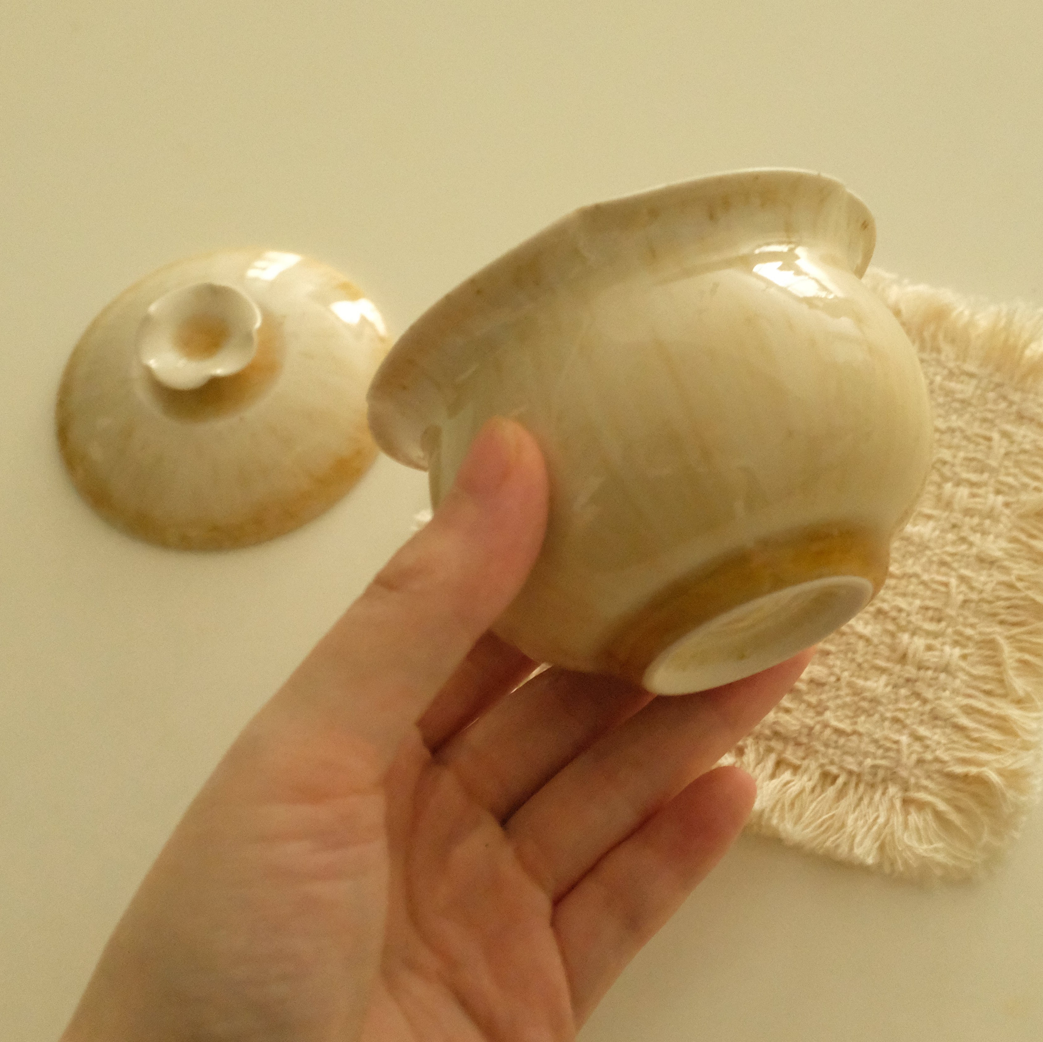 A hand holds a small beige ceramic bowl with a matching lid, with a textured beige cloth on a plain beige background.