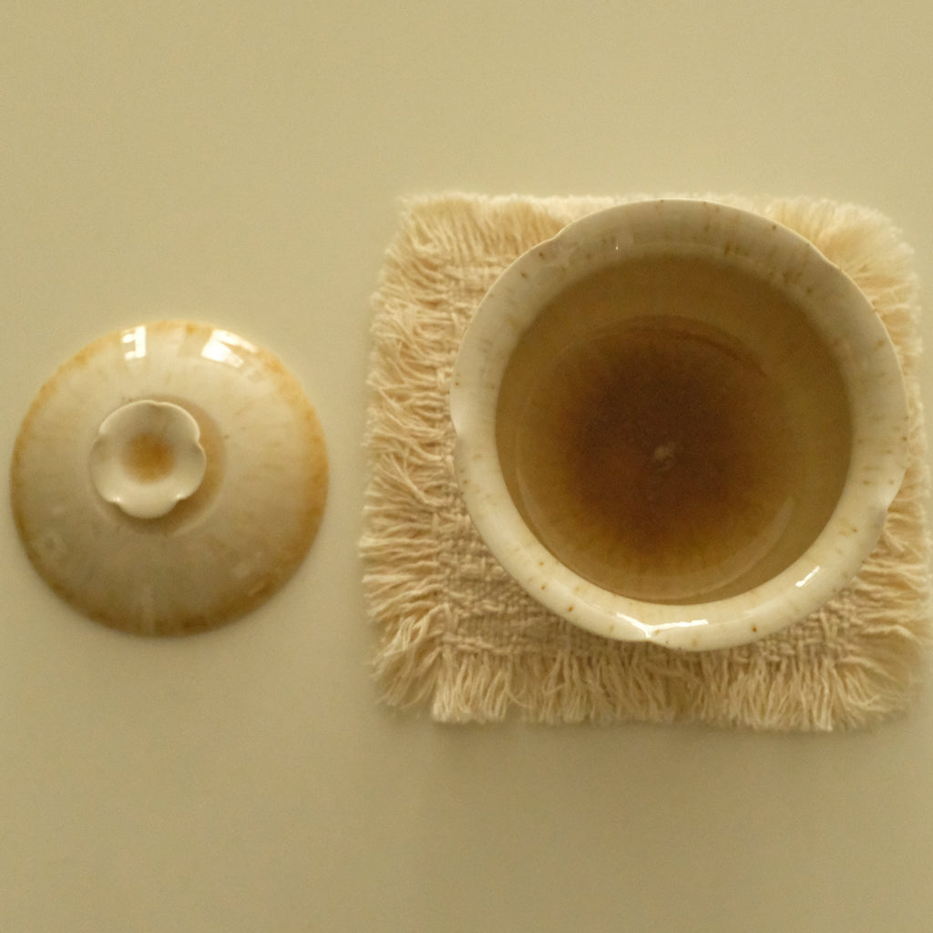 A ceramic tea cup with its lid placed beside it on a light beige background. The cup sits on a square, fringed cream coaster.
