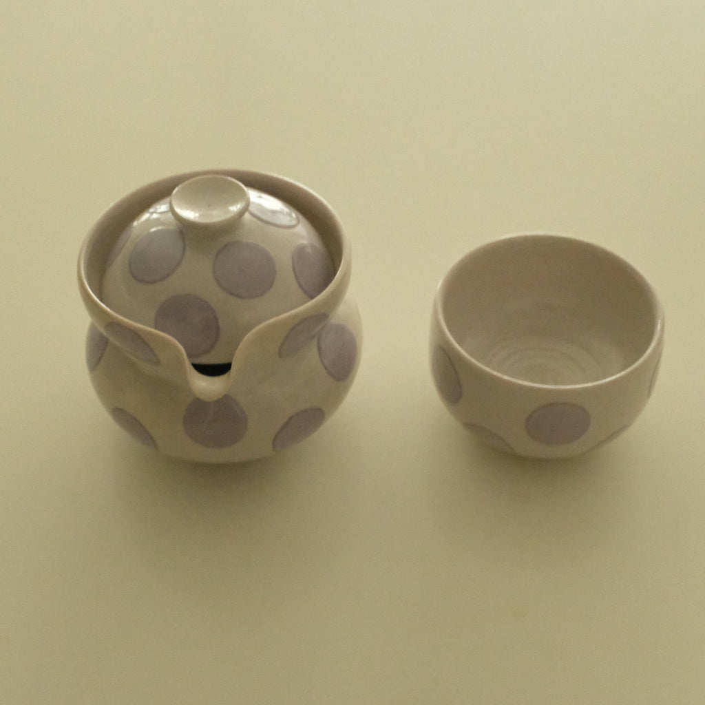 A small ceramic handless teapot with a matching small cup, both decorated with large lavender polka dots on a warm off-white background, set against a plain cream-colored surface.