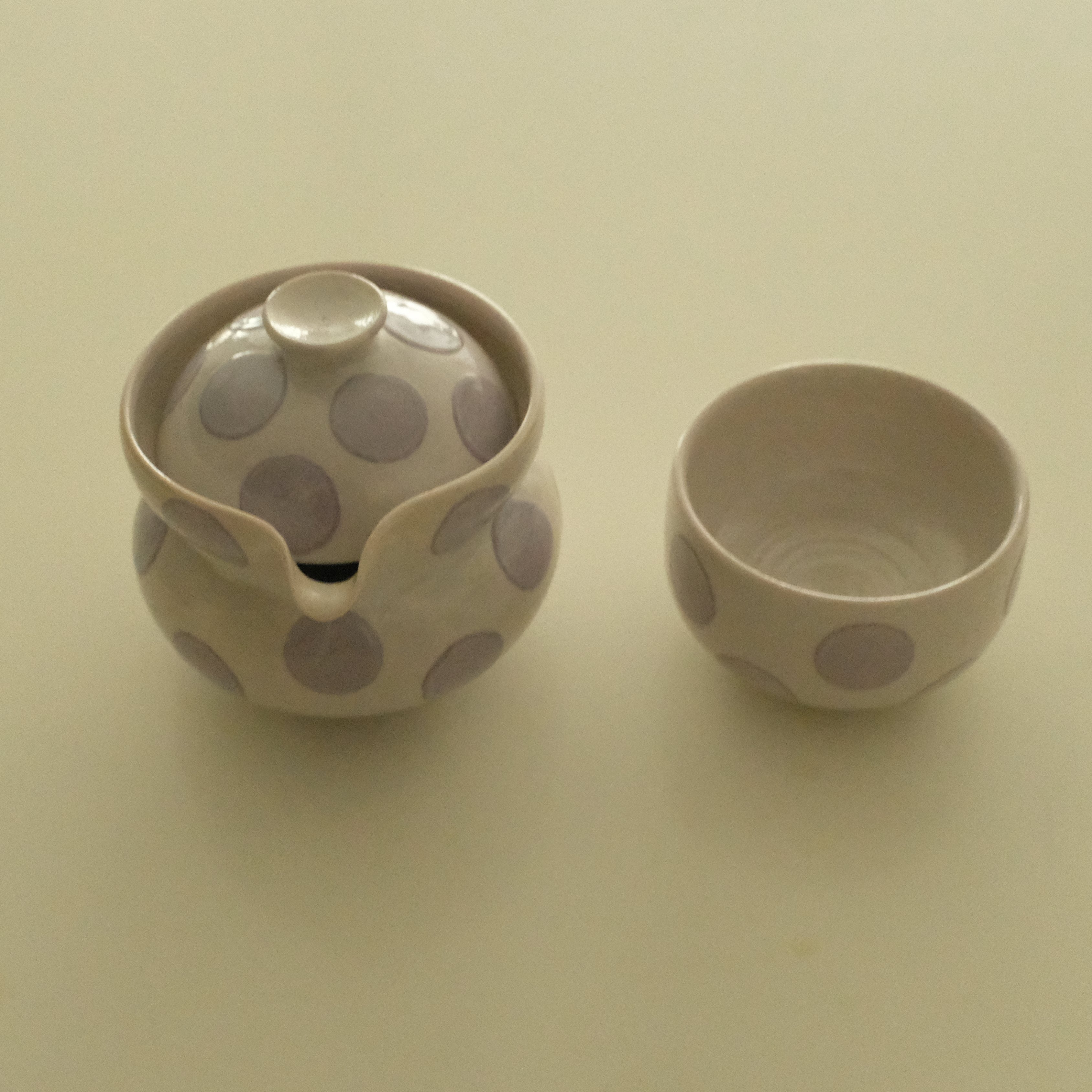 A small ceramic handless teapot with a matching small cup, both decorated with large lavender polka dots on a warm off-white background, set against a plain cream-colored surface.