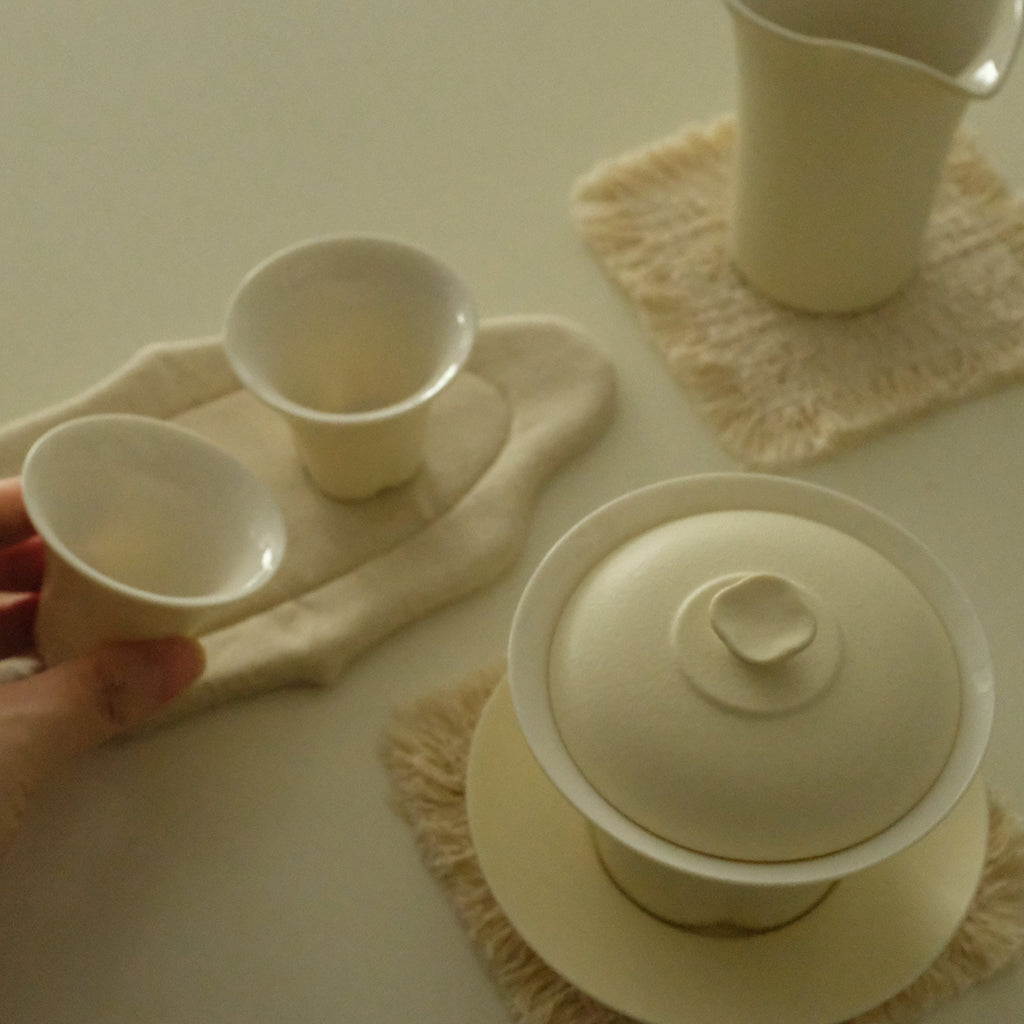 A hand holds a small ceramic cup from a tea set set on a cream-colored table. The set includes a lidded gaiwan, two small cups, a pitcher, and textured cream placemats.