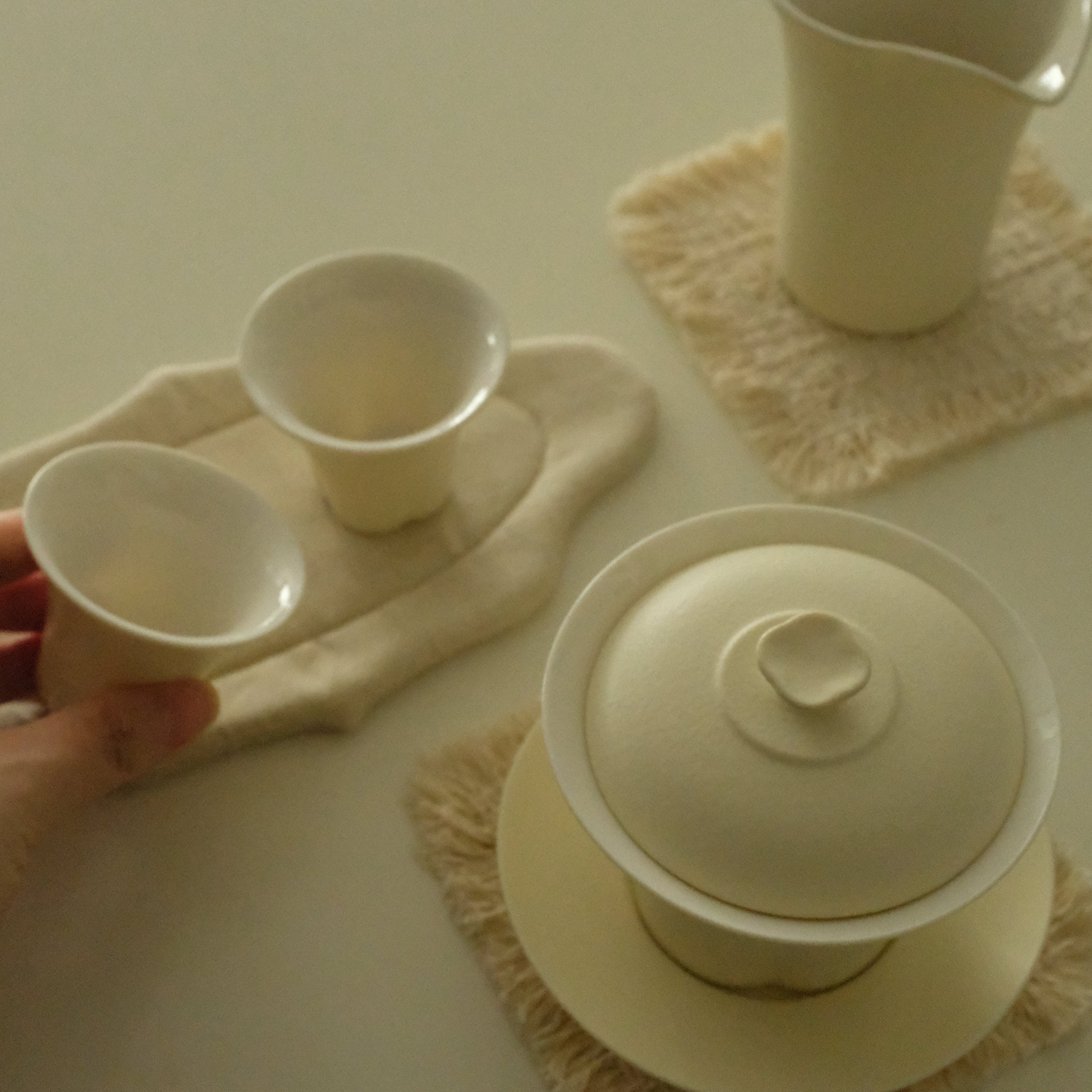 A hand holds a small ceramic cup from a tea set set on a cream-colored table. The set includes a lidded gaiwan, two small cups, a pitcher, and textured cream placemats.