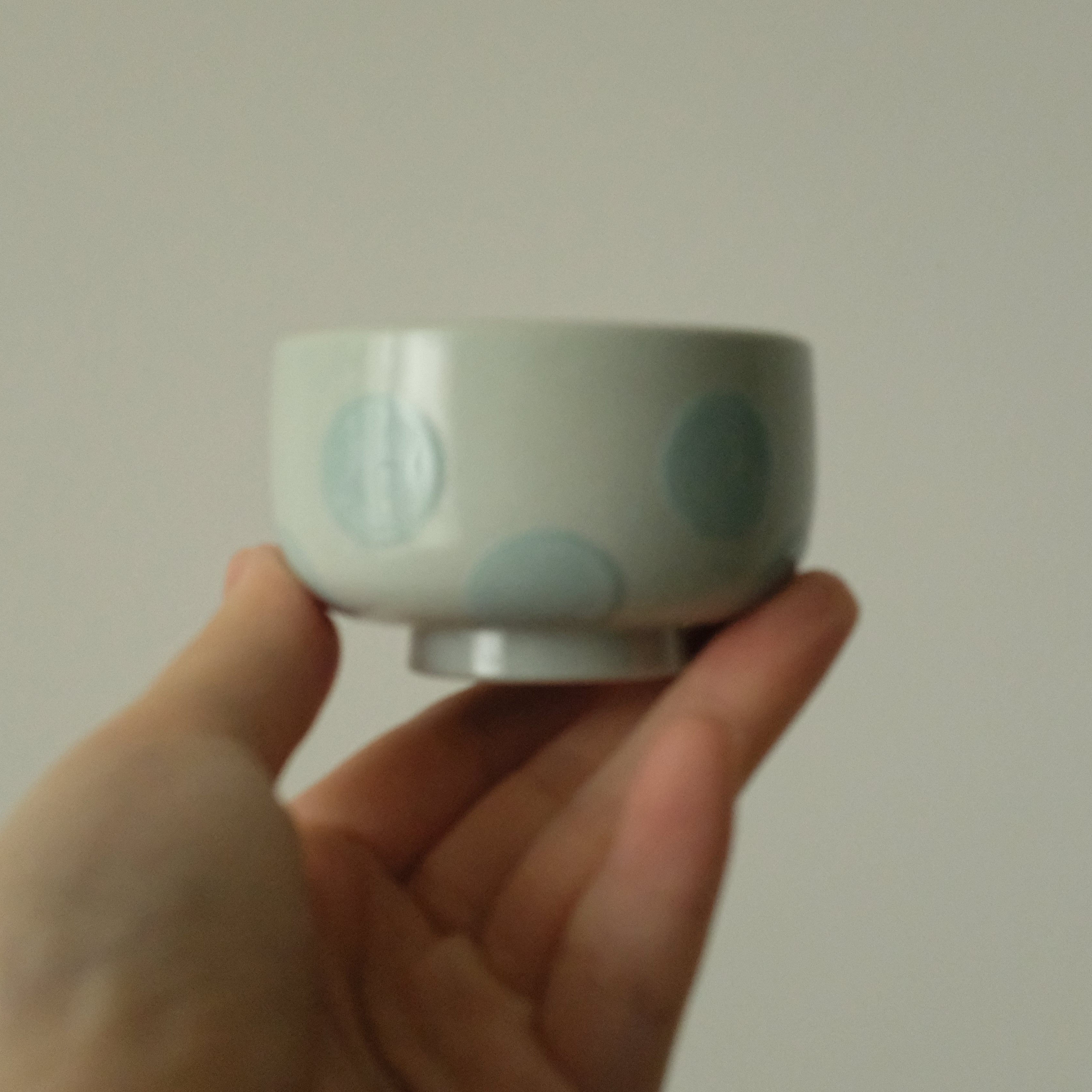 A hand holding a small white ceramic cup decorated with light blue polka dots against a plain light gray background.