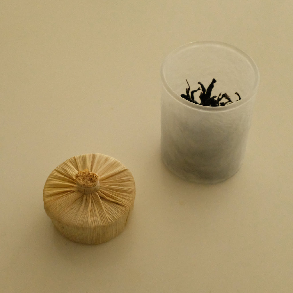 A small round beige wrapped packet sits beside a clear plastic cup containing loose dark dried leaves, photographed on a plain warm beige background.