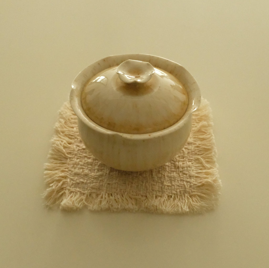 A small beige ceramic lidded cup or teacup resting on a textured cream woven coaster against a solid cream background.