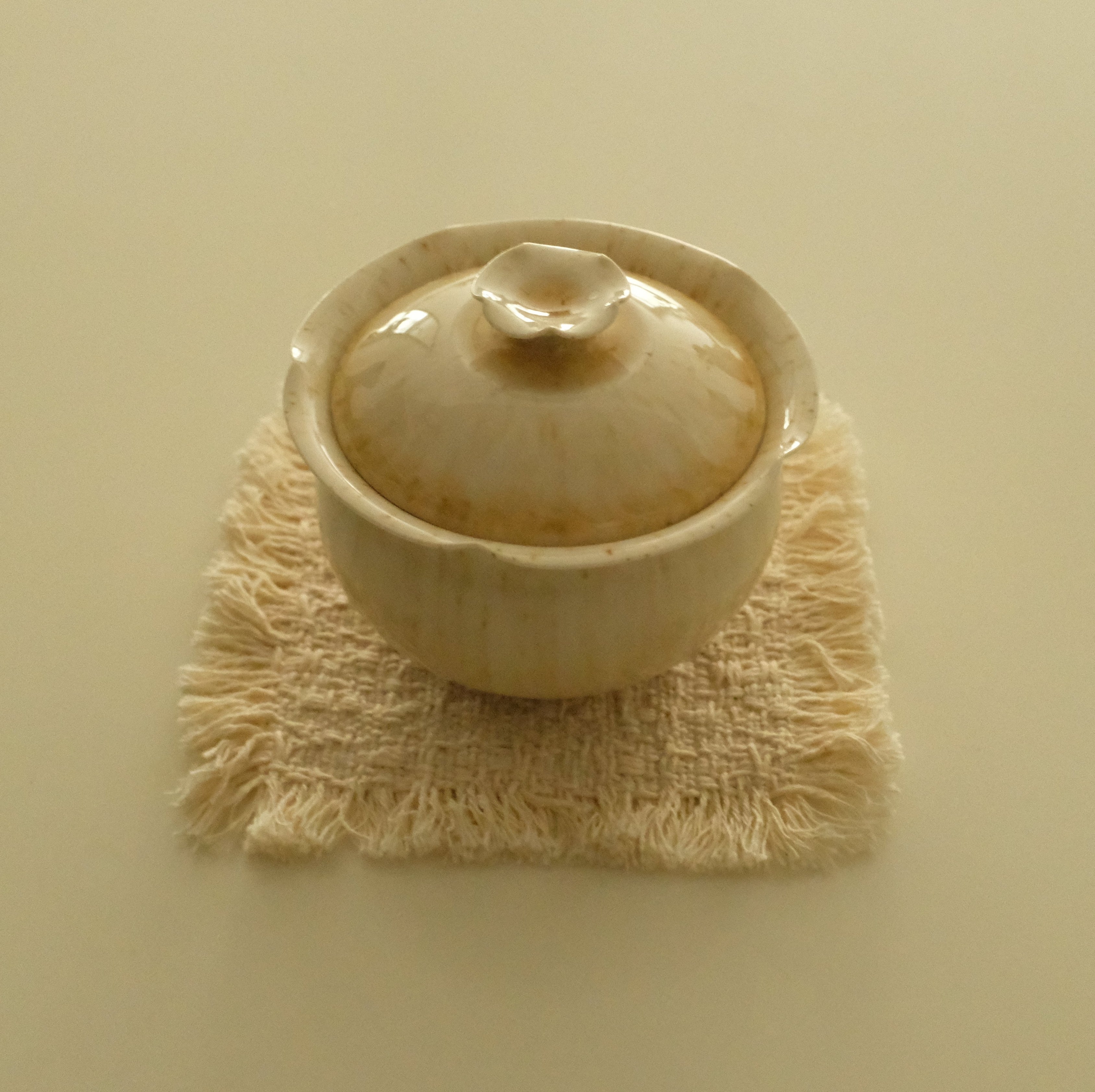 A small beige ceramic lidded cup or teacup resting on a textured cream woven coaster against a solid cream background.