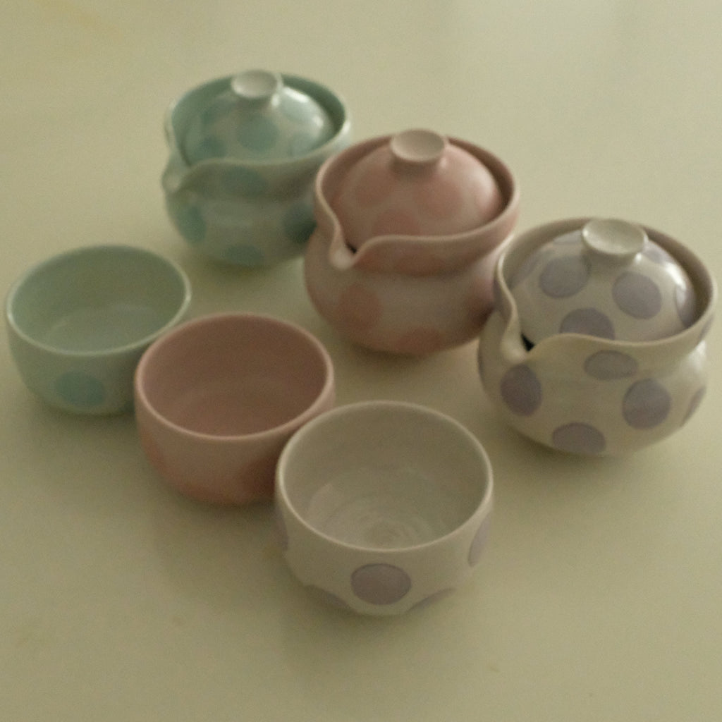 A set of six small ceramic tea cups and two lidded vessels in soft pastel colors, including ice blue, blush pink, lavender, and white with polka dots, arranged on a plain light beige surface.