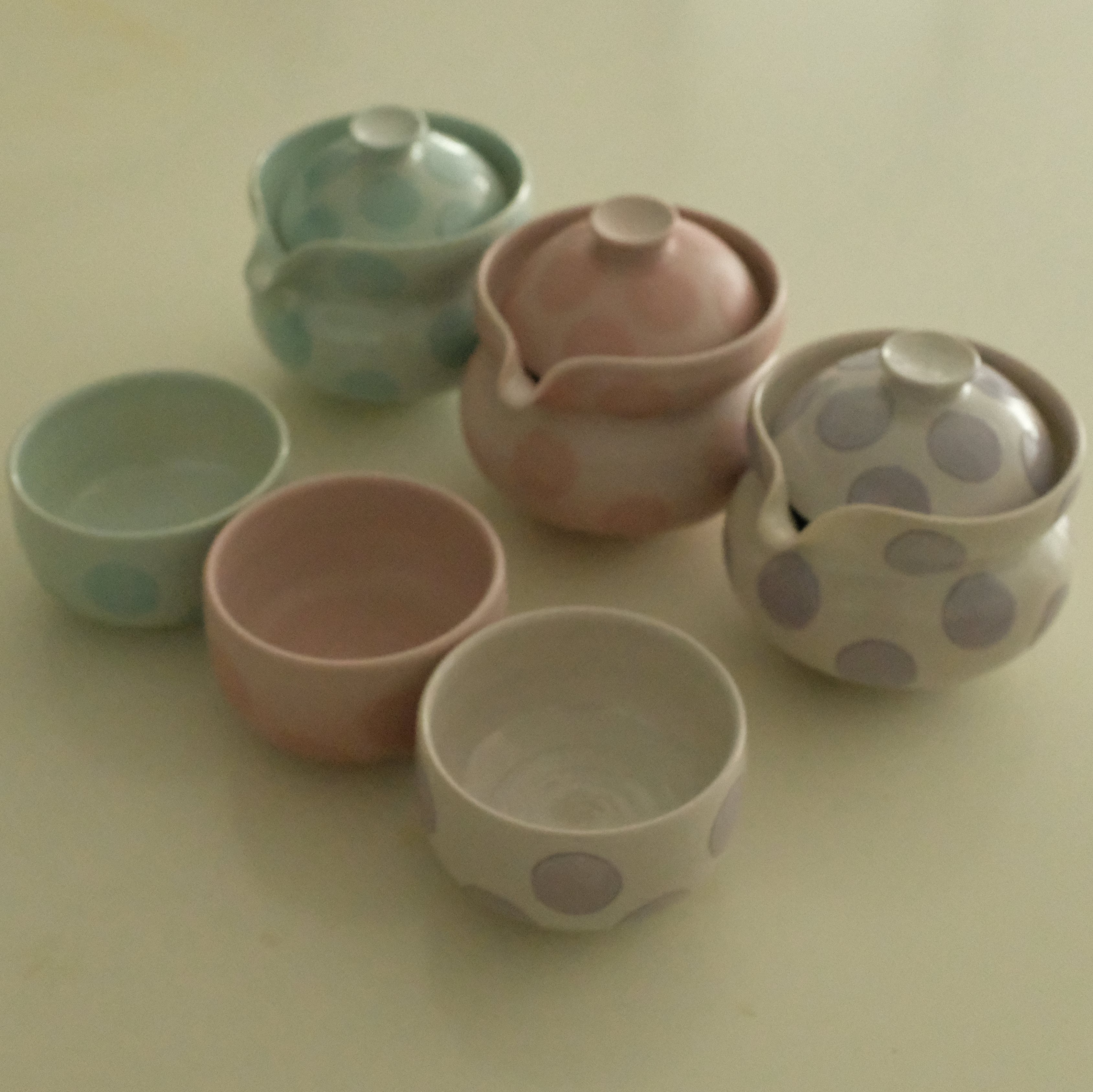 A set of six small ceramic tea cups and two lidded vessels in soft pastel colors, including ice blue, blush pink, lavender, and white with polka dots, arranged on a plain light beige surface.