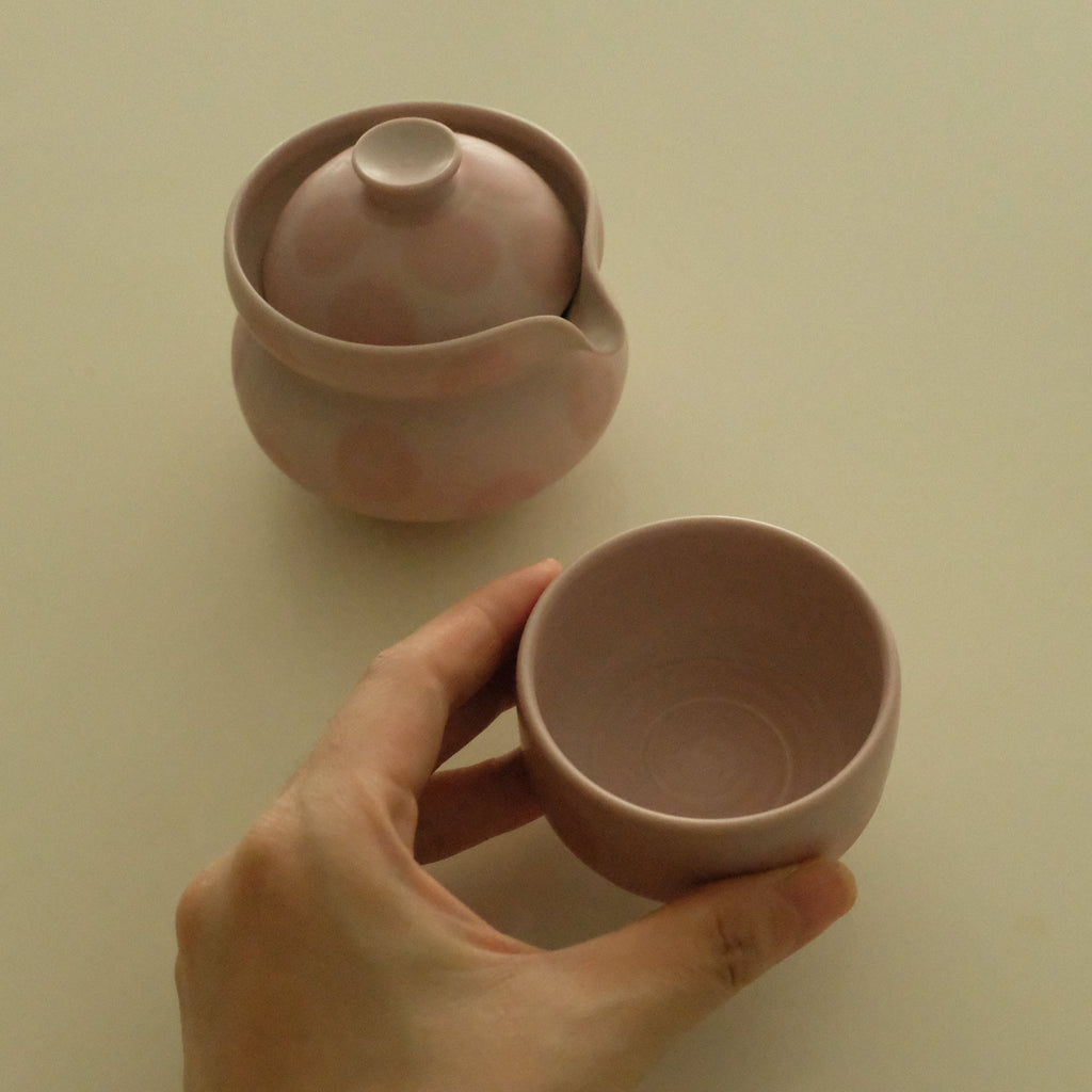 A hand holds a small pink ceramic cup while a matching pink ceramic teapot with lid sits on a plain light beige background.