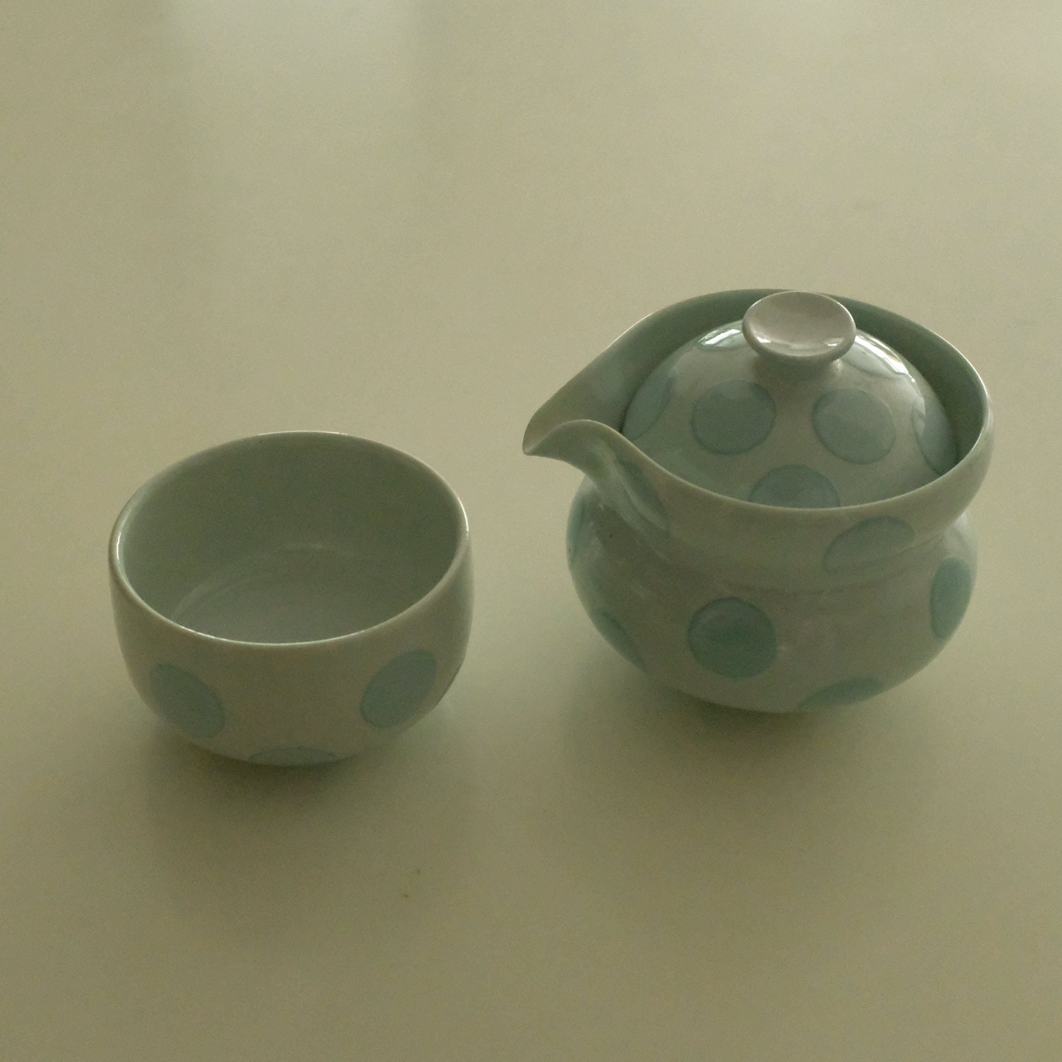 A small ceramic teapot and matching cup set in a pale mint green color with darker green polka dots, set against a plain off-white background.