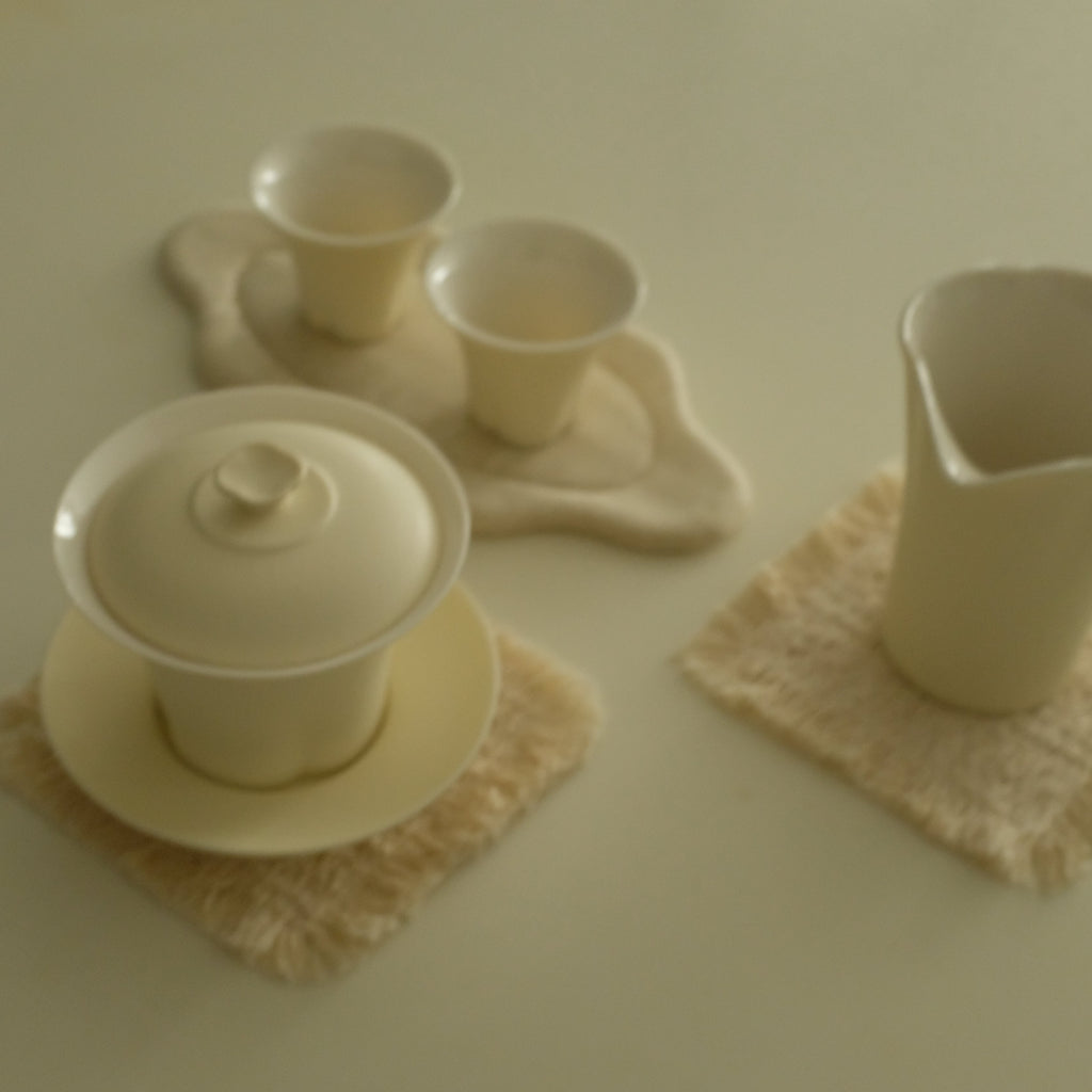 A cream-colored ceramic tea set with a lidded gaiwan, two small cups on a matching saucer, and a pitcher, arranged on textured cream-colored coasters against a solid cream background.