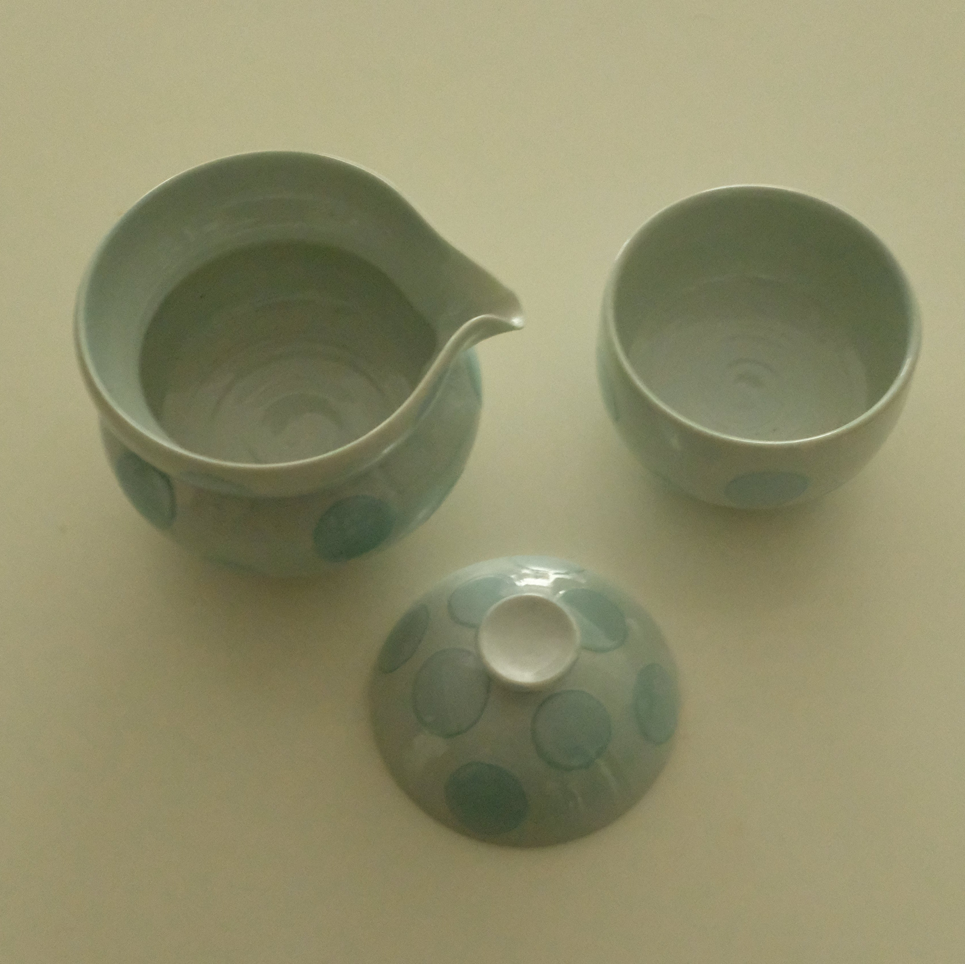 A light blue-green ceramic tea set with a pitcher, a small cup, and a matching lid, decorated with a pattern of blue-green circles on a warm cream background.