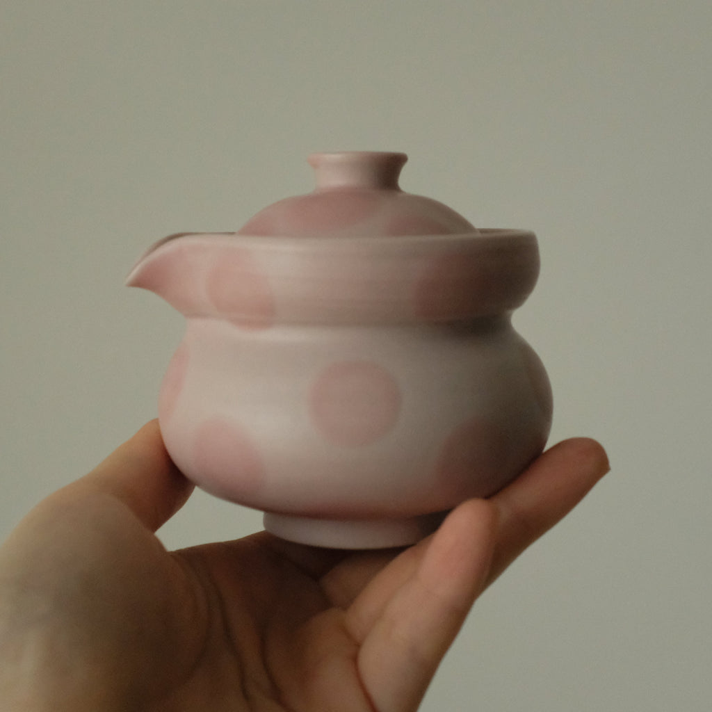 A small pink ceramic teapot with pink polka dots, held in a person's hand against a plain light gray background.