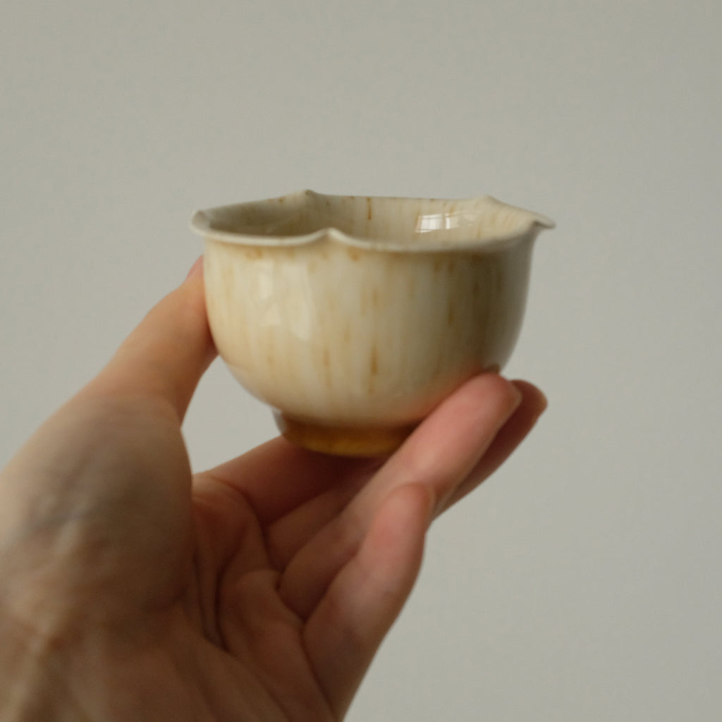 A hand holding a small ceramic cup with a scalloped rim, featuring a glossy cream glaze with subtle brown speckles and a warm tan base, set against a plain light gray background.