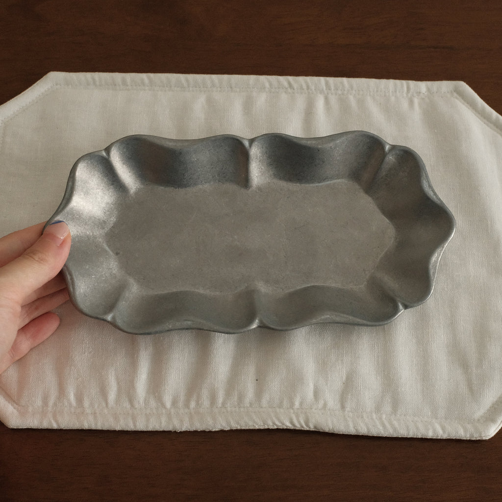 Hand holding a metal dish over a white placemat on a wooden surface