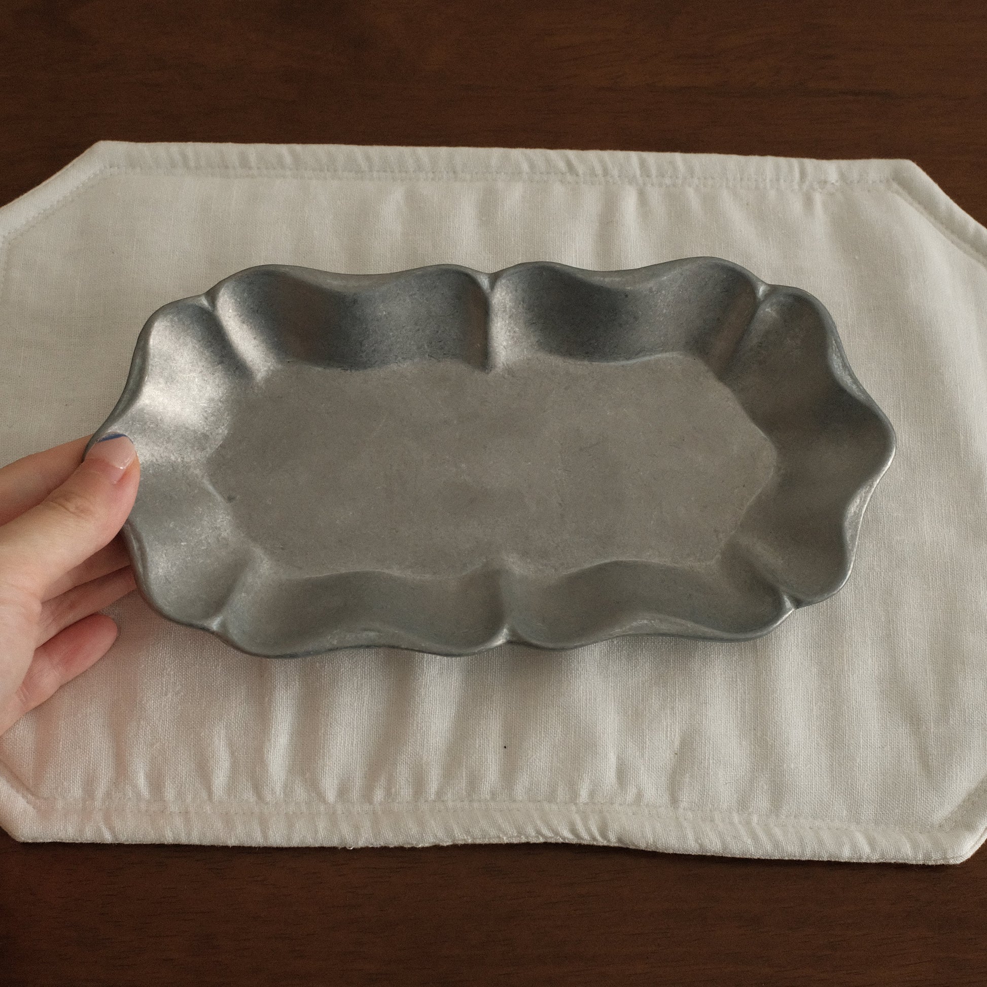 Hand holding a metal dish over a white placemat on a wooden surface