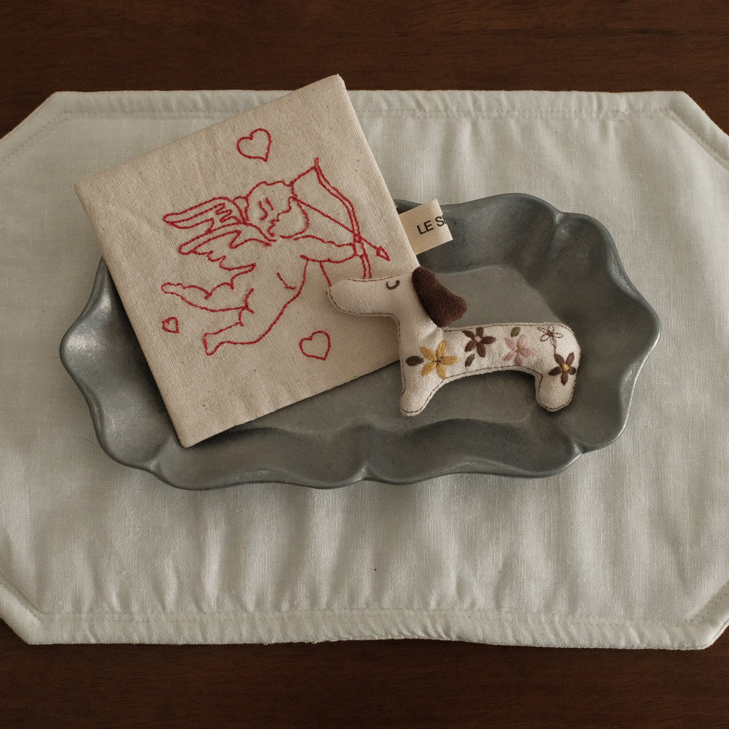 Decorative items including a small coaster with a design, a dog dachshund-shaped felt brooch, and a gray metal decorative plate on a white placemat