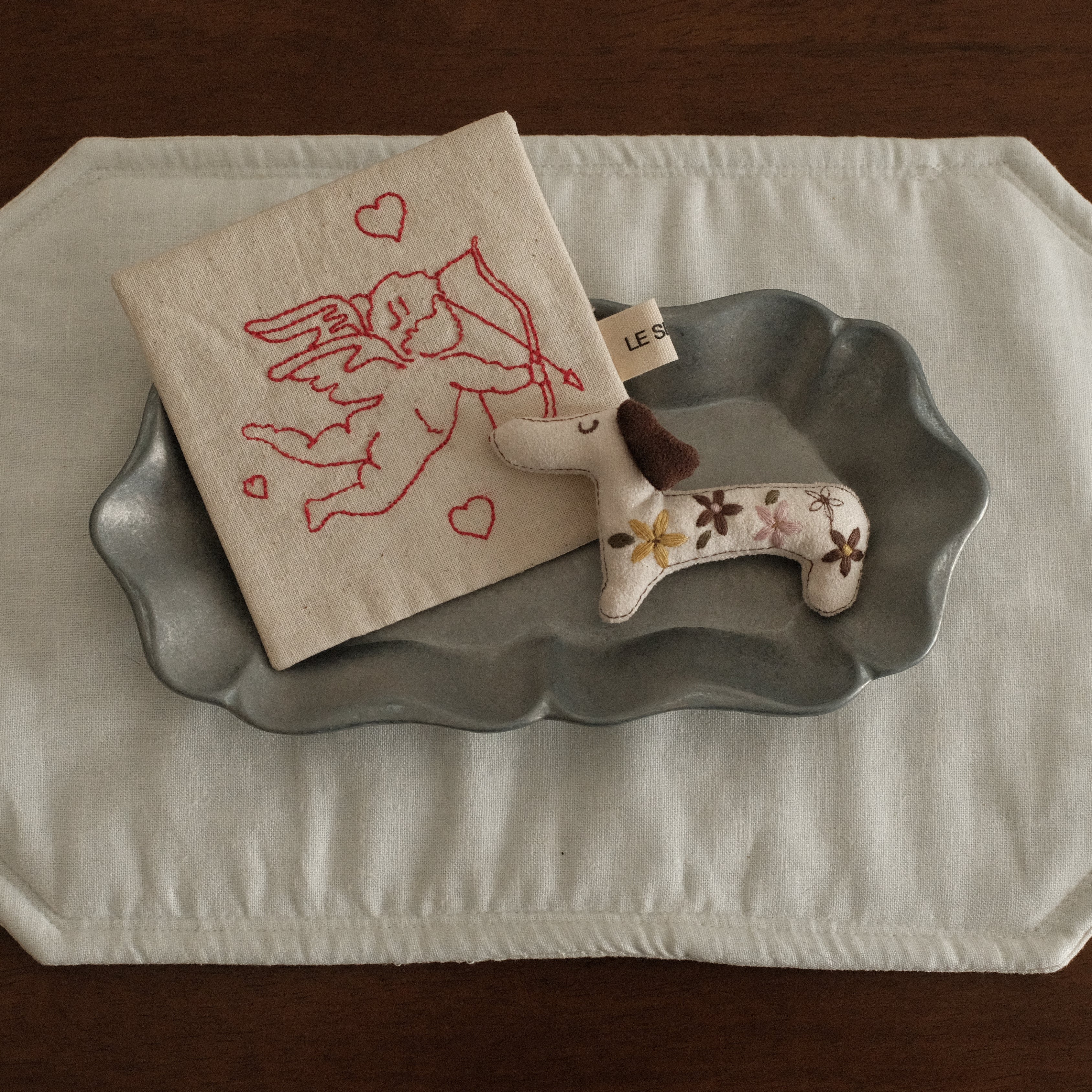 Decorative items including a small coaster with a design, a dog dachshund-shaped felt brooch, and a gray metal decorative plate on a white placemat