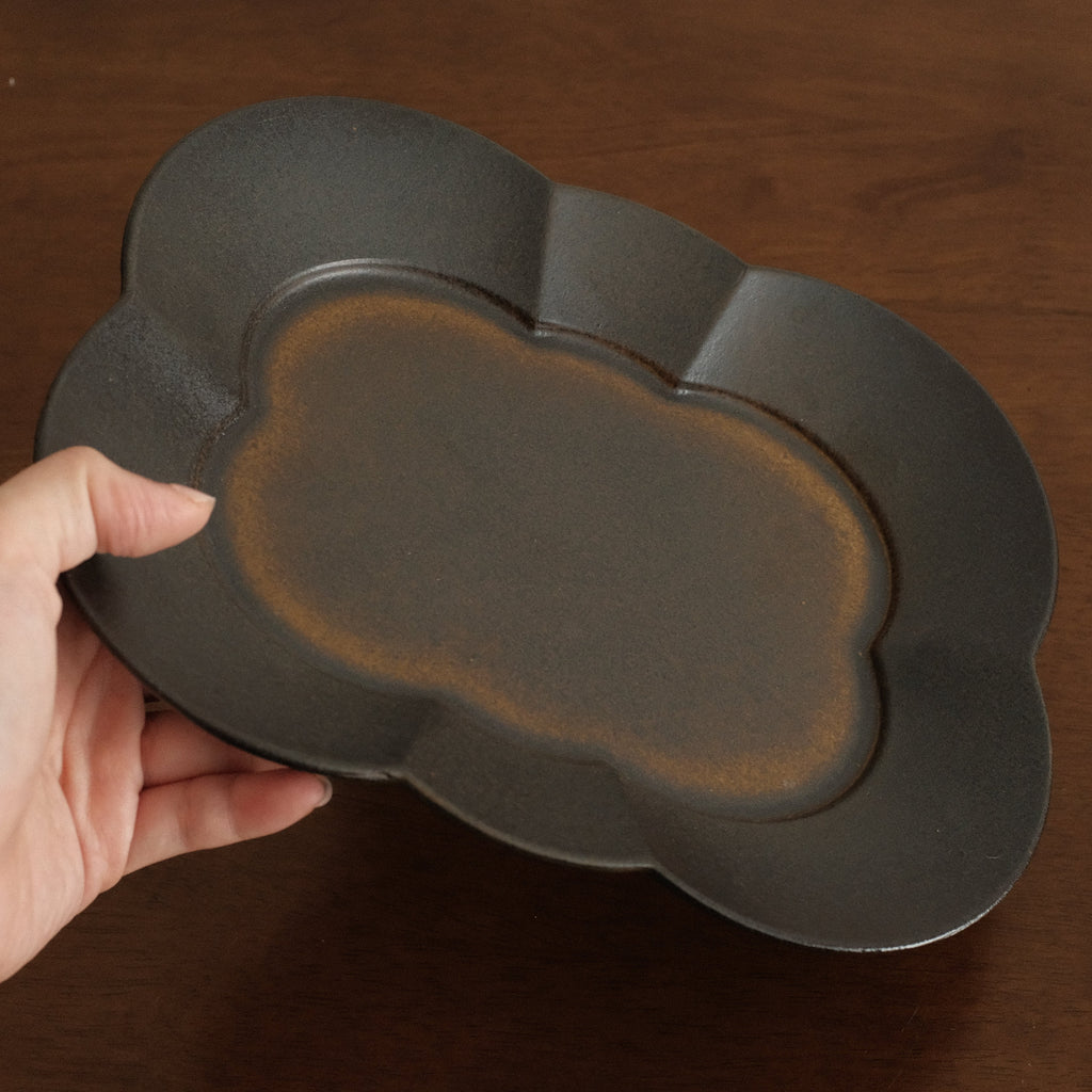 Hand holding a dark ceramic tray with a scalloped edge on a wooden surface
