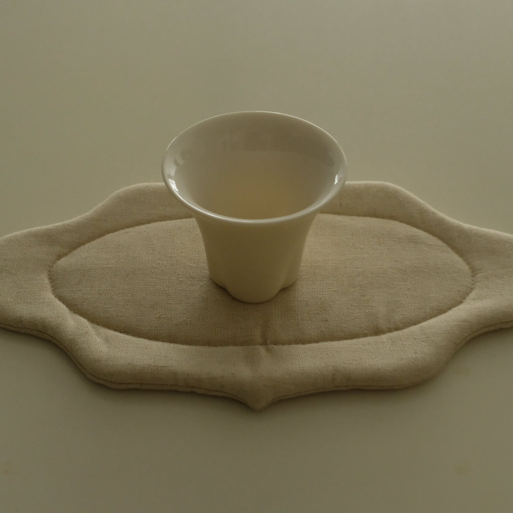 A small white ceramic cup sits centered on a beige fabric pad with an irregular scalloped edge, placed on a plain beige surface.