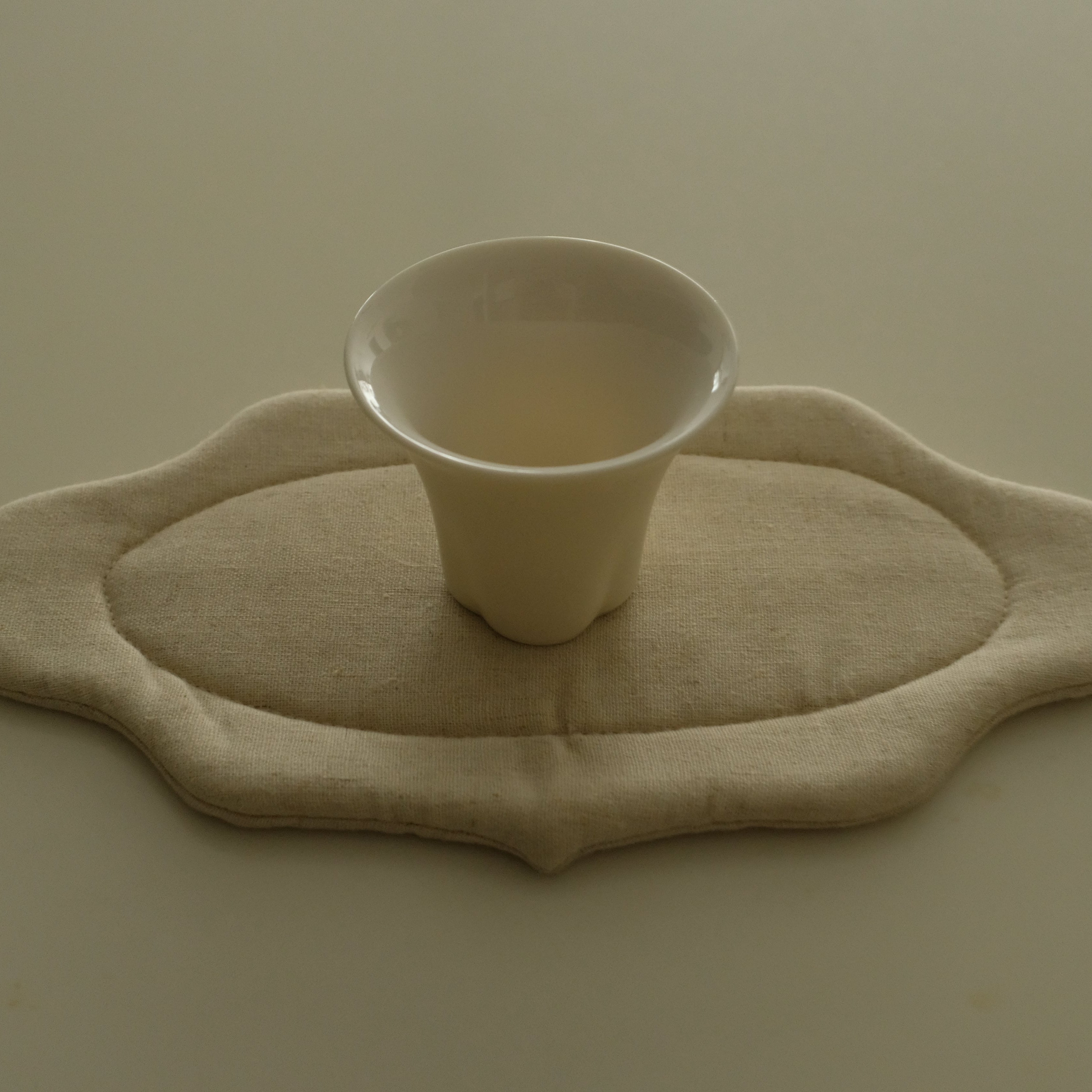 A small white ceramic cup sits centered on a beige fabric pad with an irregular scalloped edge, placed on a plain beige surface.