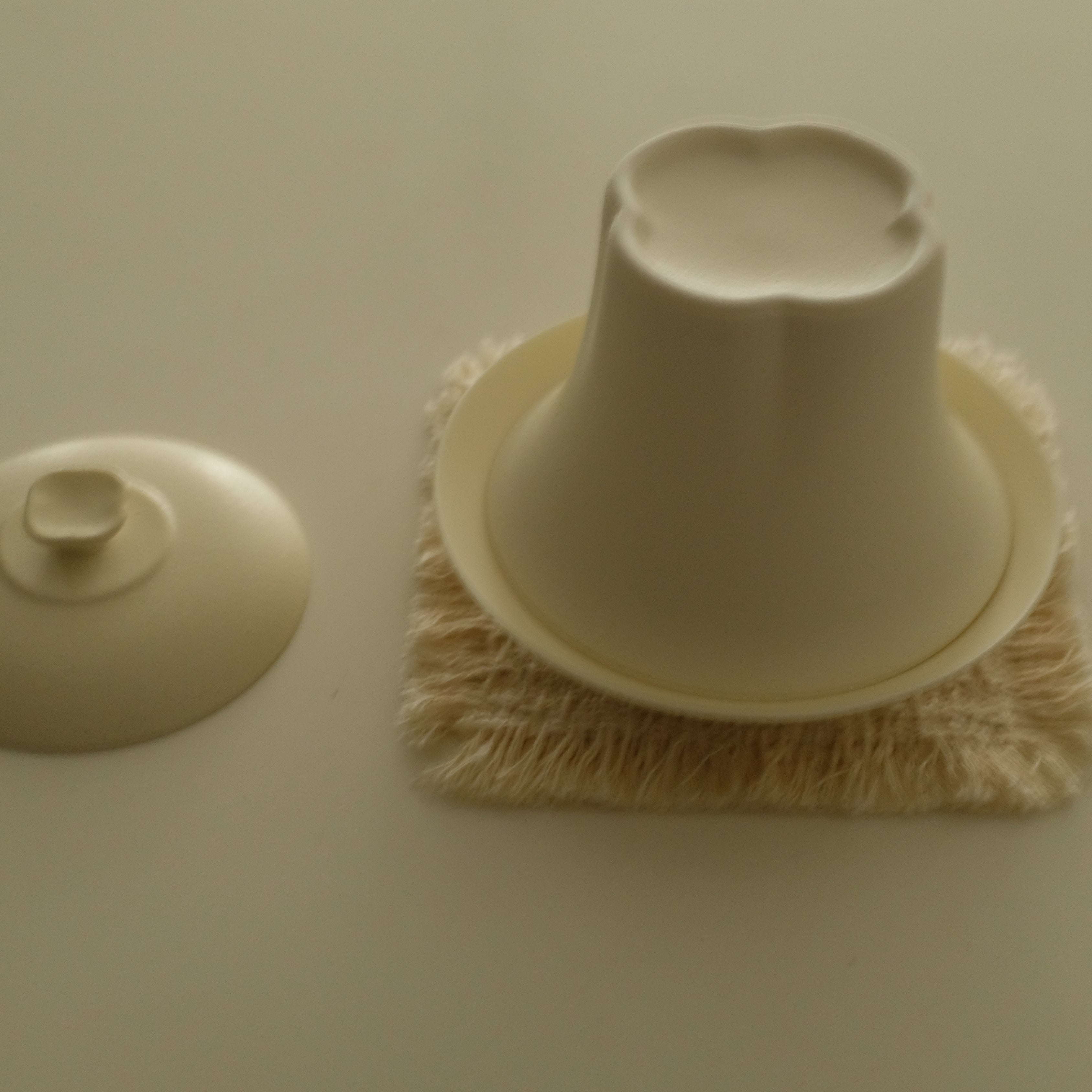 A beige ceramic or porcelain cleaning brush with a conical bristle head resting on a square beige pad, shown next to its matching beige lid on a plain beige background.