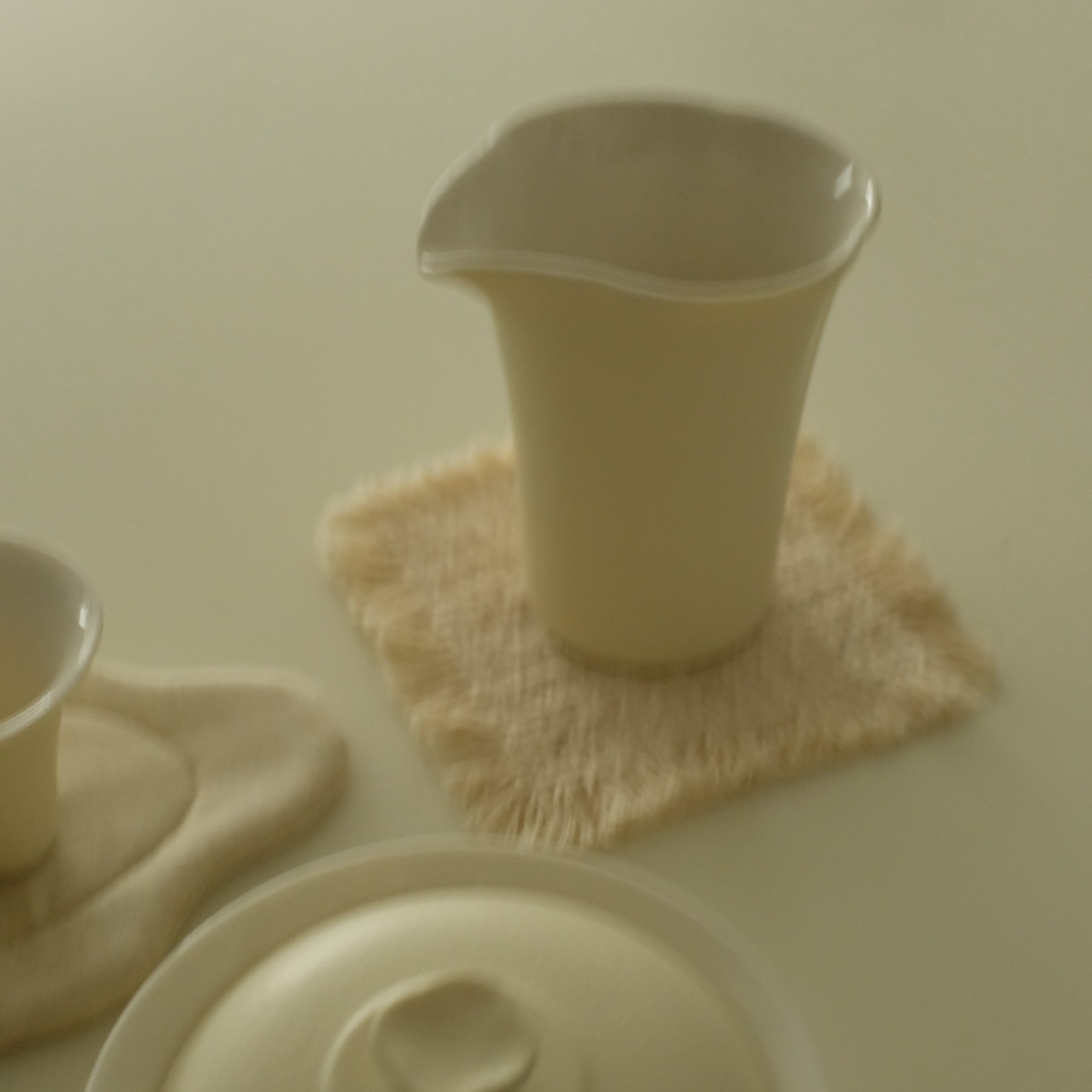 A set of matte ceramic tea or coffee vessels on a warm beige surface, including a tall pitcher, two small cups, a lid, and a woven coaster.